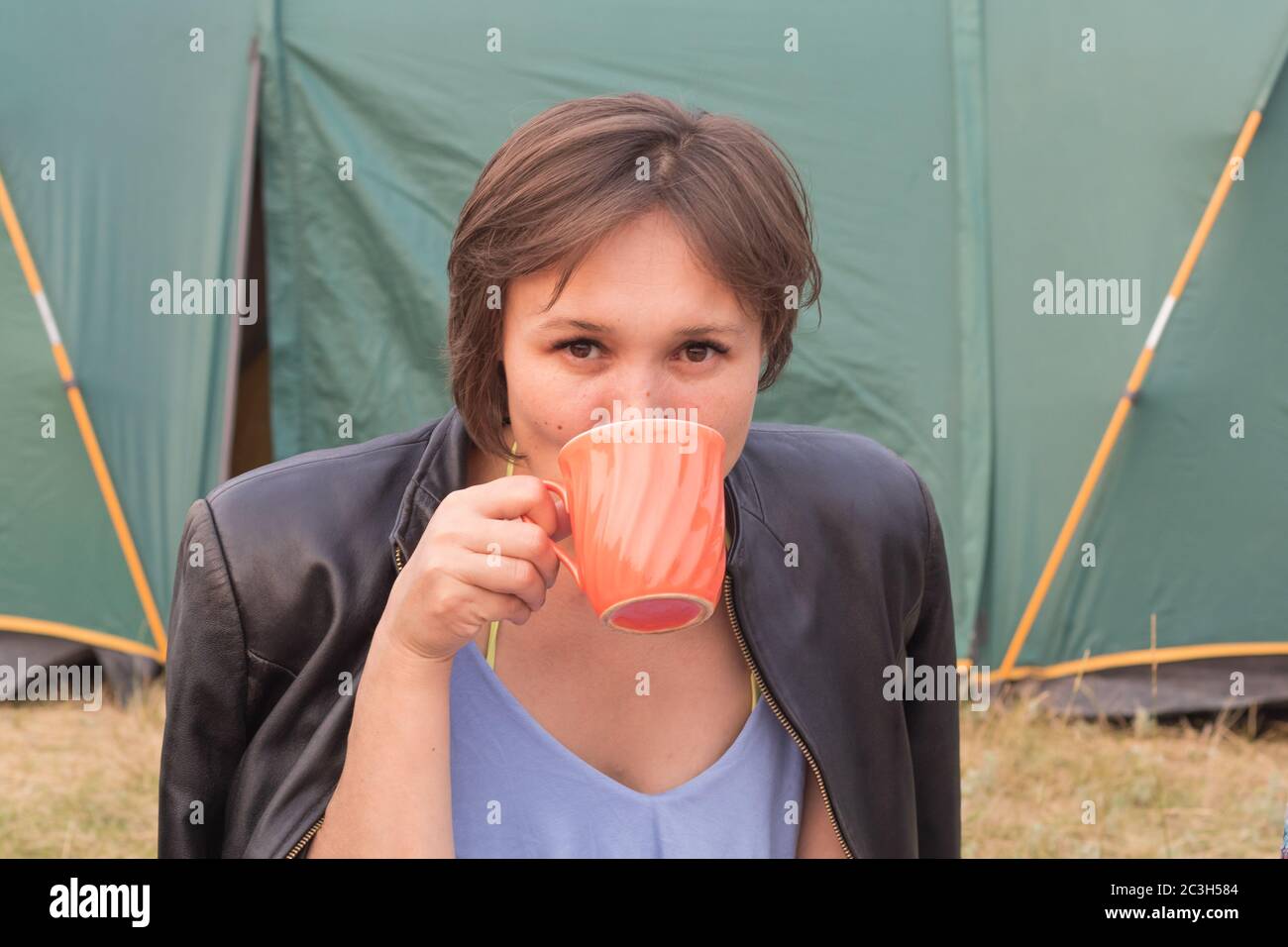 Hübsche junge Frau in einem Top und Jacke, Tee aus einem großen Becher trinken und auf die Kamera auf dem Hintergrund des Zeltes schauen. Inlandstourismus concep Stockfoto