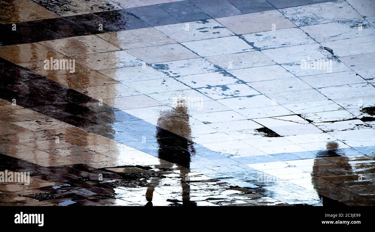 Verschwommene Spiegelschatten Silhouette von Menschen, die an einem regnerischen Tag auf einer feuchten Straßenpfütze in der Fußgängerzone der Stadt spazieren Stockfoto