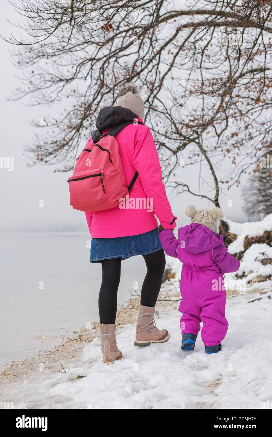 Blick von hinten auf eine Mutter und ihre Tochter, die auf Schnee am Wintersee spazieren gehen. Stockfoto