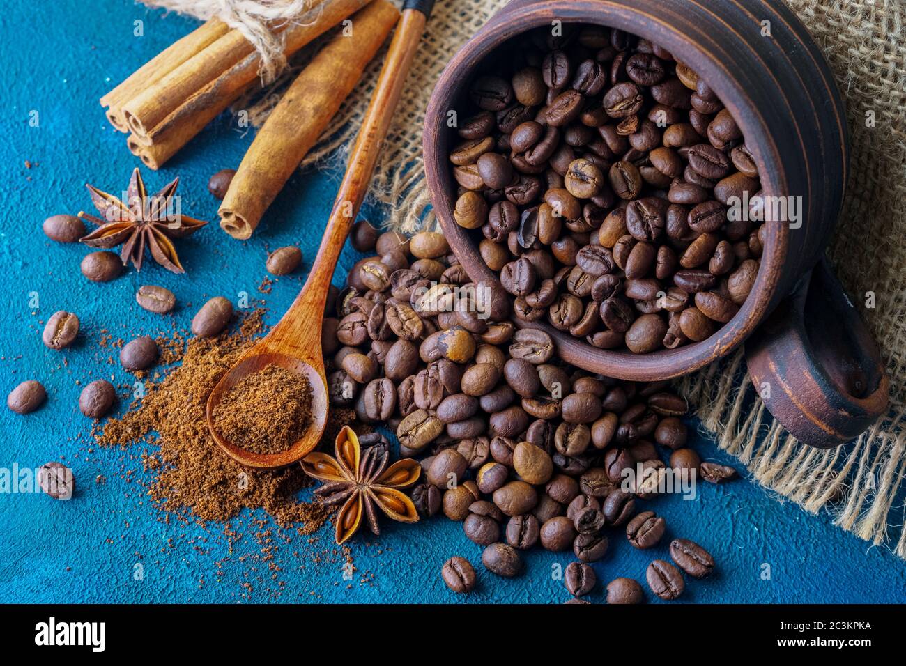 Kaffeesorten, die aus einer Tonschale ausströmen und auf einem blauen texturellen Hintergrund verstreut sind, Anissterne, Zimtstangen und gemahlener Kaffee in einem Holzlöffel Stockfoto