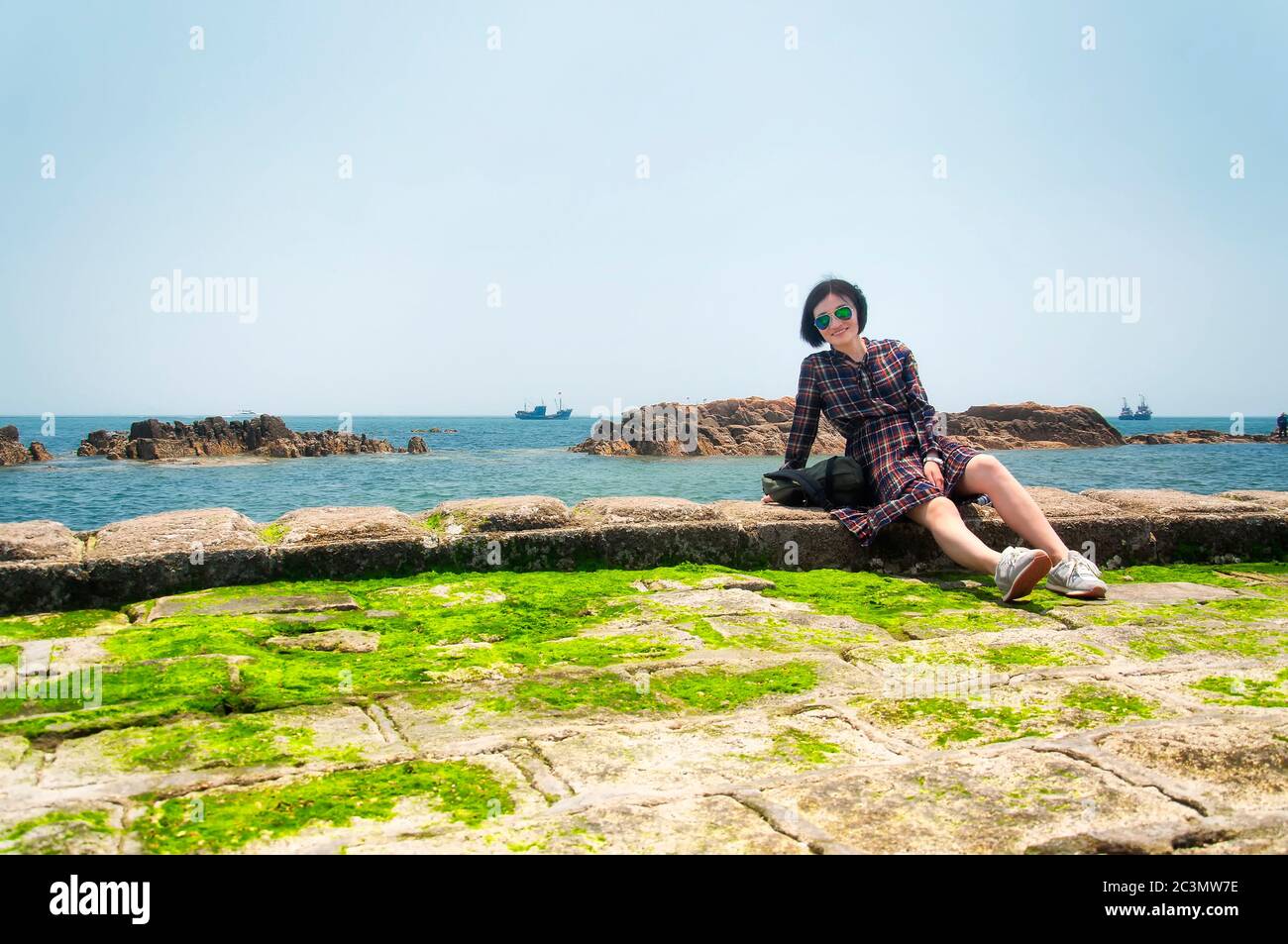 Eine glückliche chinesin, die an einem sonnigen Tag in der Nähe der felsigen Küste der Fushan Bay in der chinesischen Provinz qingdao shandong sitzt. Stockfoto