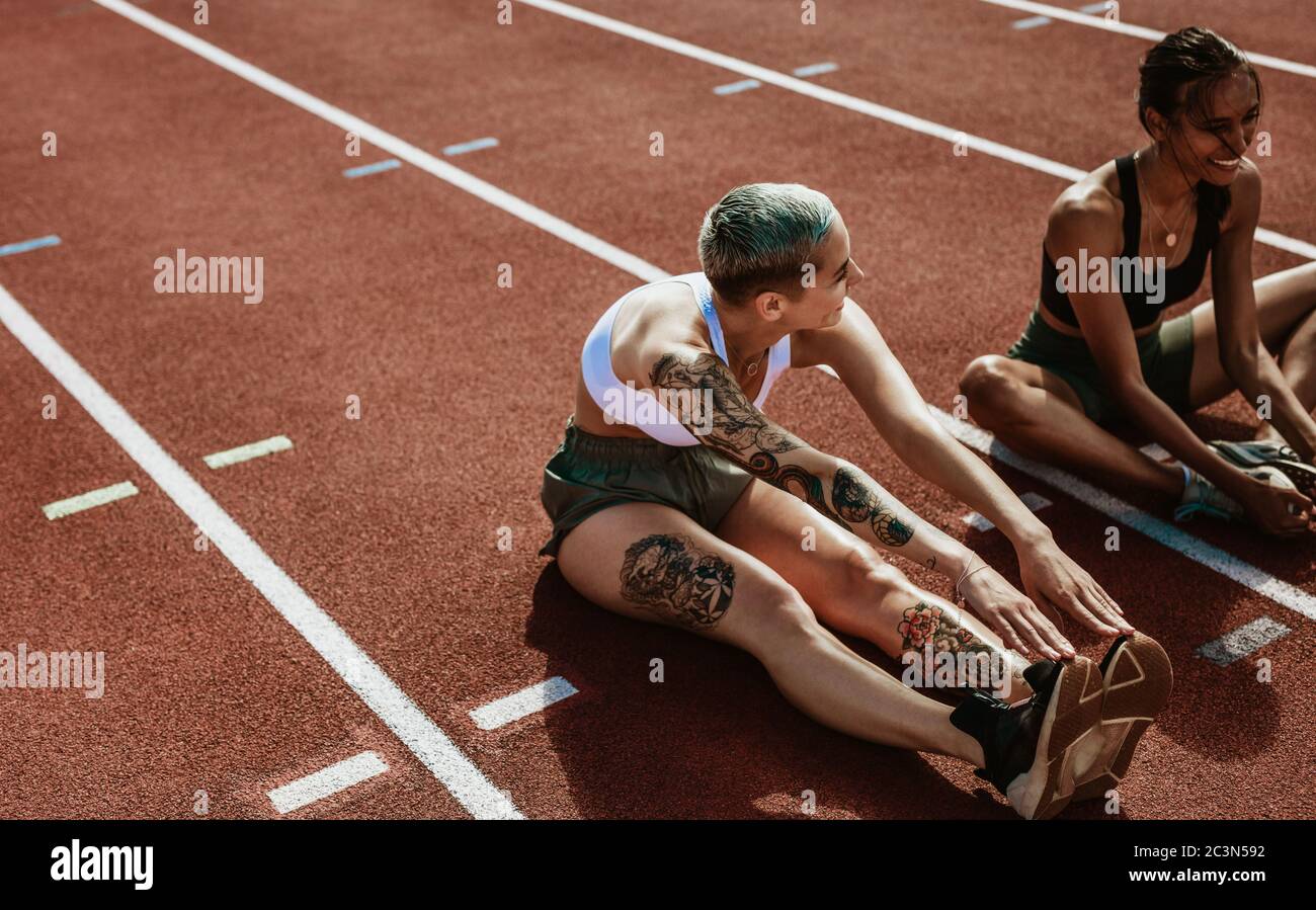 Zwei weibliche Athleten machen Dehnübungen sitzen auf Laufstrecke. Läuferin sitzt auf der Laufstrecke und streckt die Beine. Stockfoto