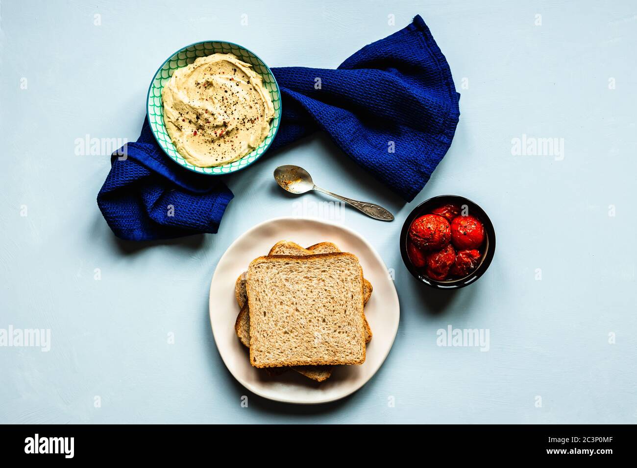 Gebackene Tomaten, Hummus und Brot. Ein tiefblaues Handtuch und ein Löffel. Blauer Hintergrund. Horizontales Bild von oben. Stockfoto