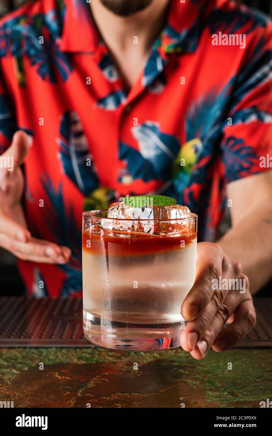 Barkeeper serviert dem Gast ein Getränk. Cocktail in einem Steinglas mit kristallklarem Eis, garniert mit einem Minzblatt. Farbenfroher Tiki-Stil. Stockfoto