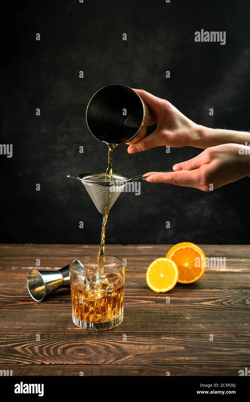 Einen Cocktail pruefen, ihn aus einem Shaker durch ein Sieb in ein mit Eiswürfeln gefülltes Steinglas gießen. Schneiden Sie orange und eine Jigger im Hintergrund Stockfoto