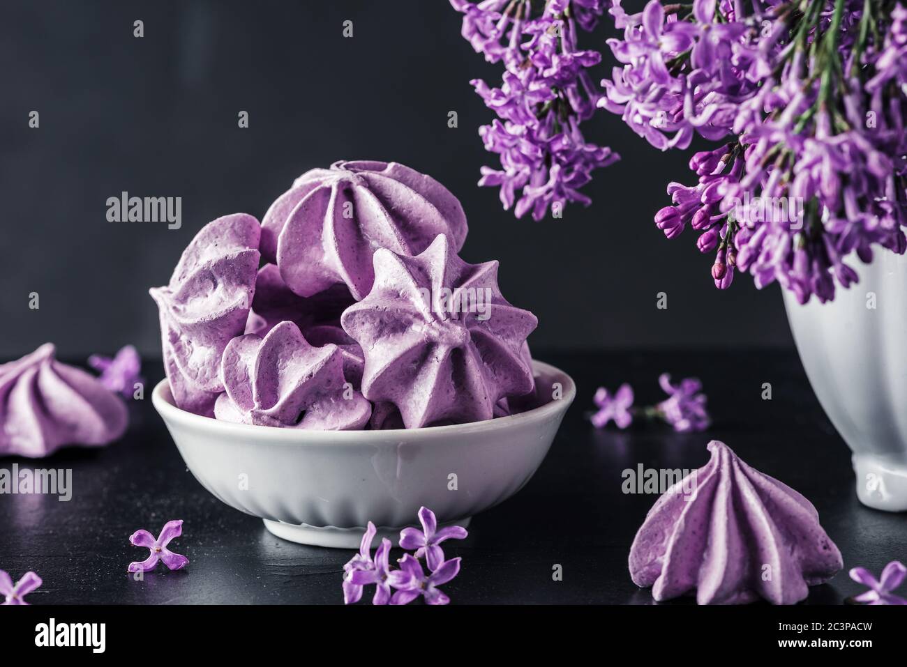 Lila Meringue Cookies mit lila Blumen in Vase auf dunklem Hintergrund. Low-Key. Stillleben. Künstlerische Aufnahme Stockfoto