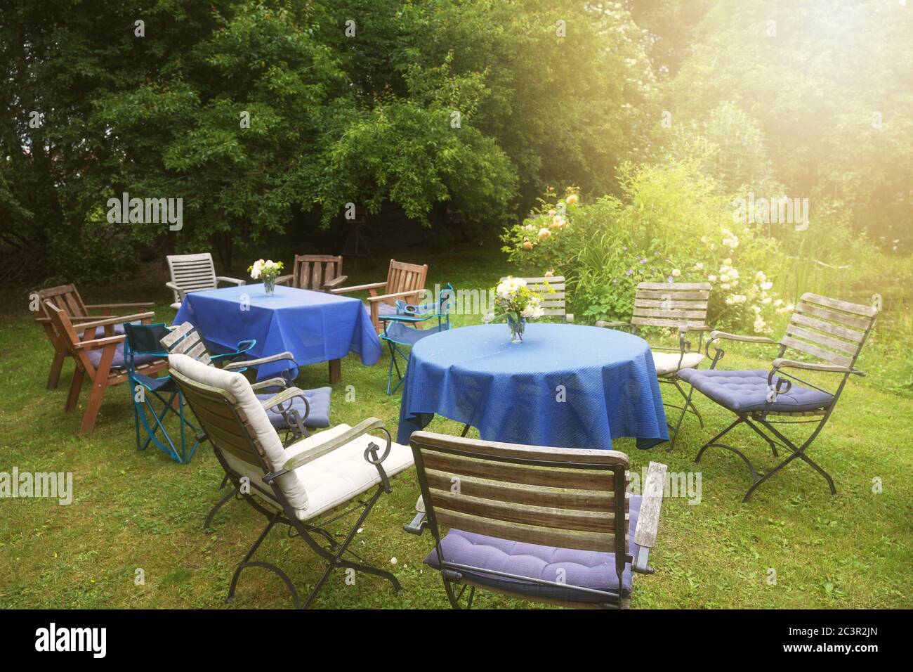 Richten Sie sich für eine private Gartenparty mit Tischen, blauen Tischdecken und vielen verschiedenen Stühlen in einem ländlichen Hinterhof in goldenen späten Nachmittag Sonnenlicht, selec Stockfoto
