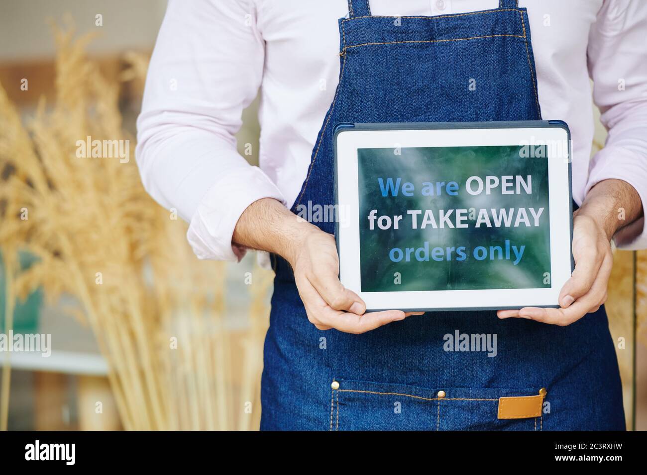 Bäckereibesitzer in Denim Schürze zeigt Tablet-Computer mit wir sind offen für Take Away Bestellungen nur Inschrift Stockfoto