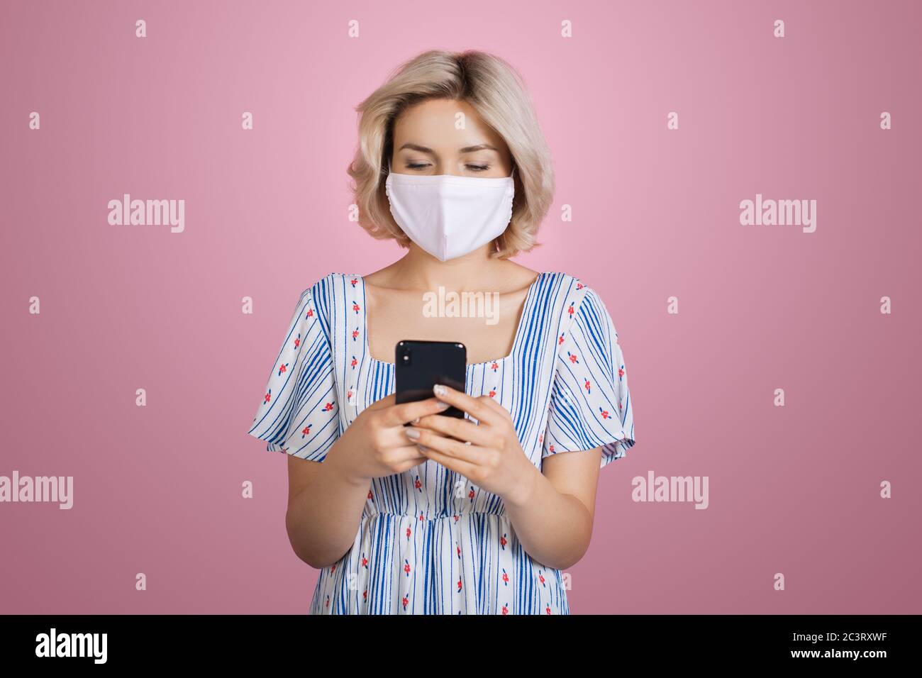 Schöne blonde Frau in medizinischen Maske trägt ein schönes Kleid ist auf Handy posiert auf einer rosa Studio Wand chatten Stockfoto