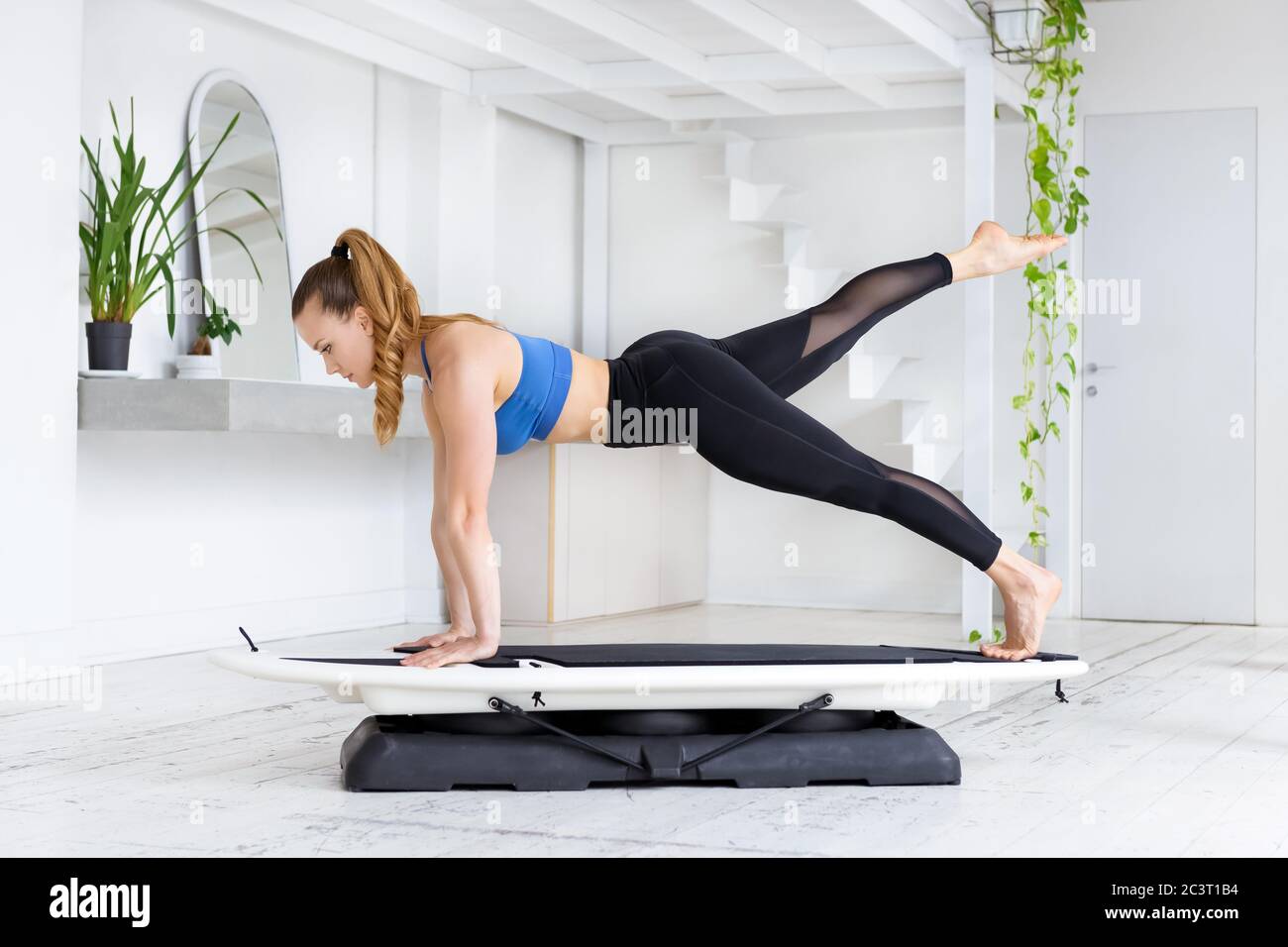 Sportlich fit junge Frau tut eine fit Surf-Plank Kick back Yoga Pose auf einem Brett in einer Seitenansicht in einem High-Key-Fitnessstudio mit frischen grünen Pflanzen in einem Gesundheits-und Stockfoto