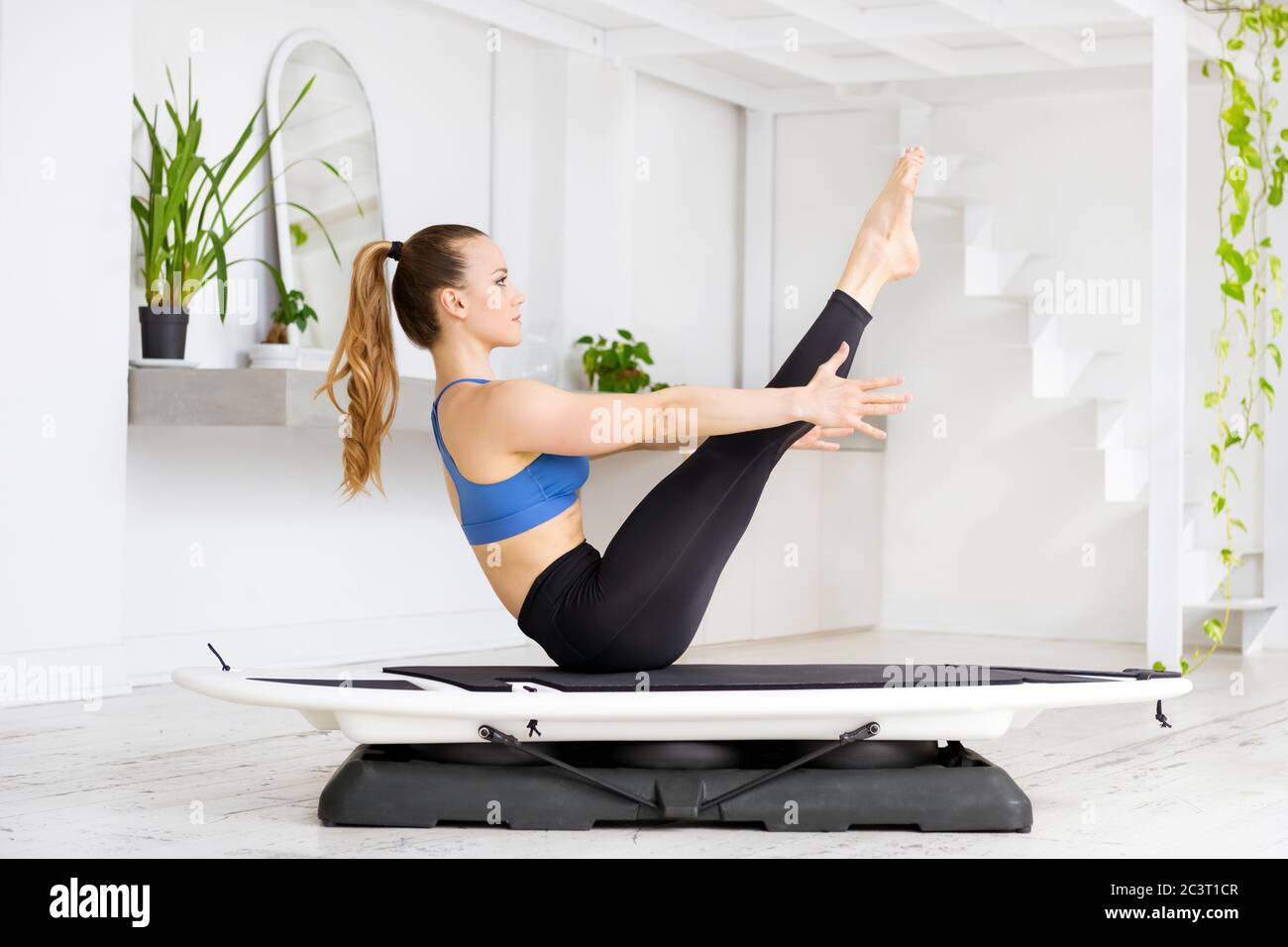 Sportlich fit junge Frau tut ein Surfen Yoga Boot gerade Beine Pose auf einem Fitness-Surfer in Seitenansicht in einem High-Key-Fitnessstudio mit grünen Pflanzen in einer Gesundheit Stockfoto