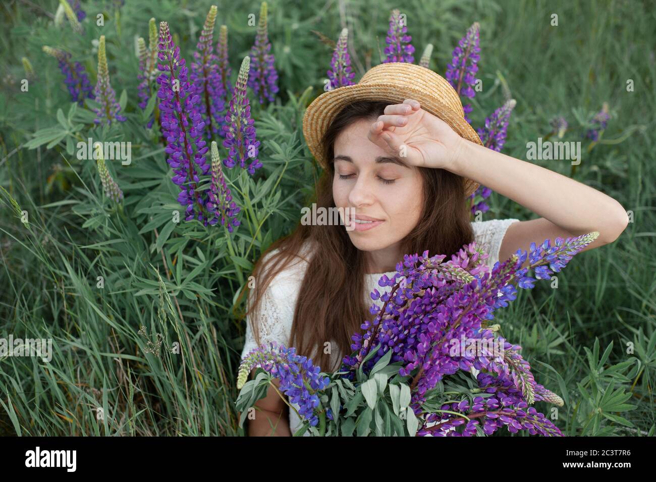 Schöne romantische Frau mit Blumenstrauß Lupinen lächelt freudig in weißem Kleid und Hut sitzt im Feld der lila Lupine Blumen. Weicher selektiver Fokus Stockfoto