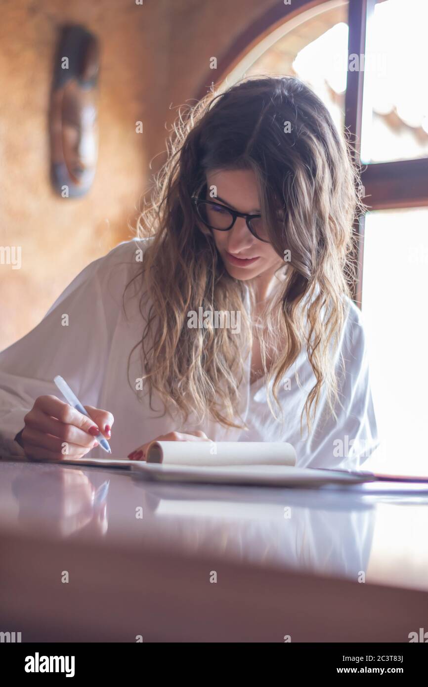 Junge Brünette Frau mit Brille und lockeren Haaren Schreiben in einem Notebook am Schreibtisch sitzen. Stockfoto
