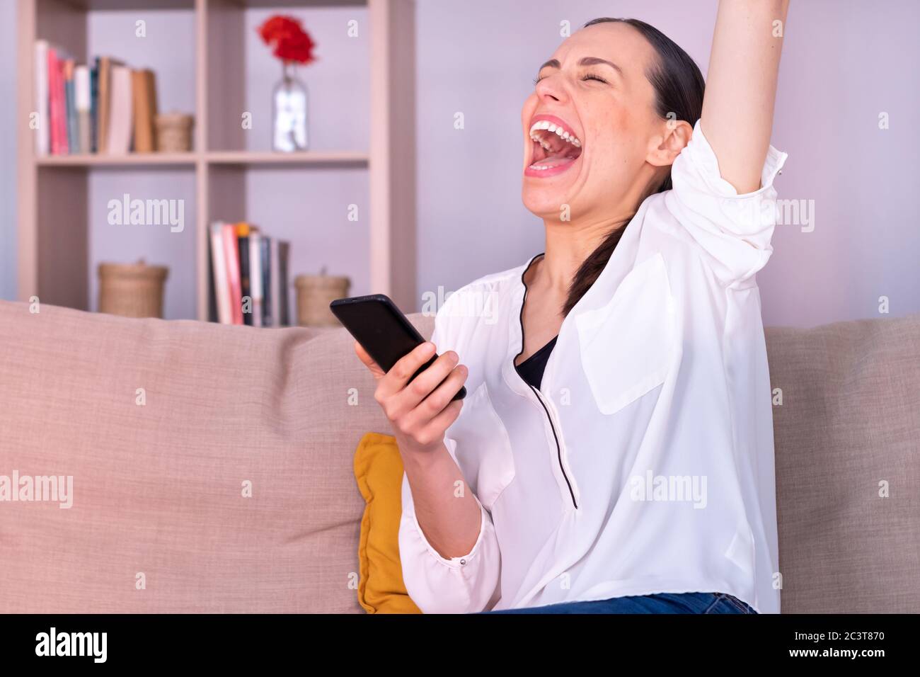 Schöne junge Brünette Frau euphorisch erhält gute Nachrichten durch das Smartphone sitzen auf dem Sofa zu Hause. Sie trägt ein weißes Hemd und einen Pferdeschwanz. Stockfoto