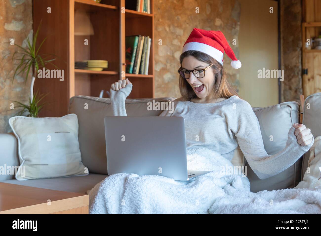 Euphorische Frau lächelt mit ausgestreckten Armen Brille und Weihnachtsmann Hut sitzt auf dem Sofa zu Hause vor einem Laptop Stockfoto