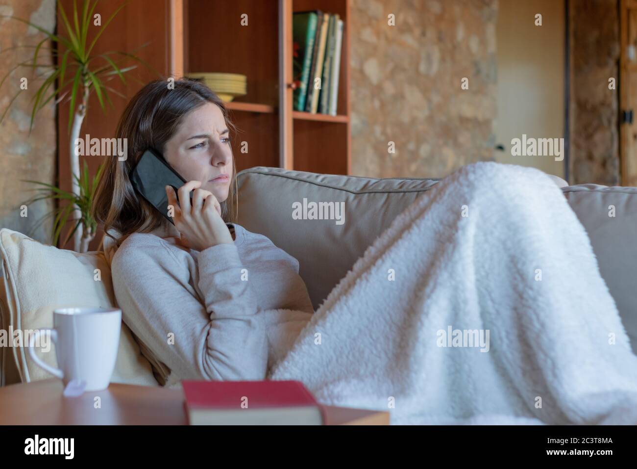 Junge ziemlich ernst Brünette Frau im Gespräch auf dem Smartphone sitzen auf dem Sofa zu Hause mit einer Decke bedeckt Blick zum Horizont. Stockfoto
