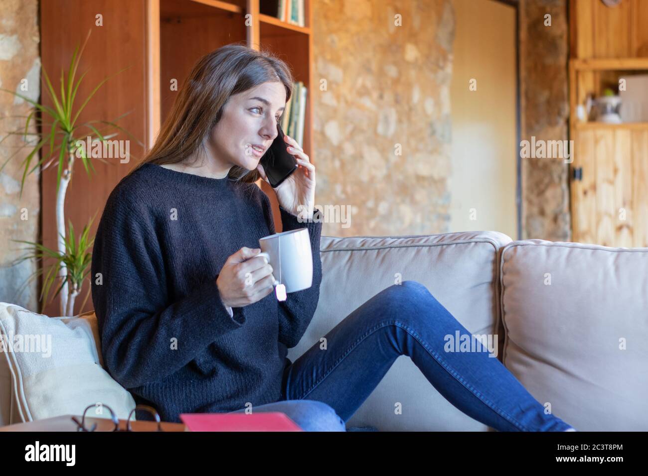 Seriöse junge Frau im Gespräch auf dem Handy sitzen auf dem Sofa zu Hause trinken einen Tee in schwarzen Pullover und blaue Jeans Stockfoto