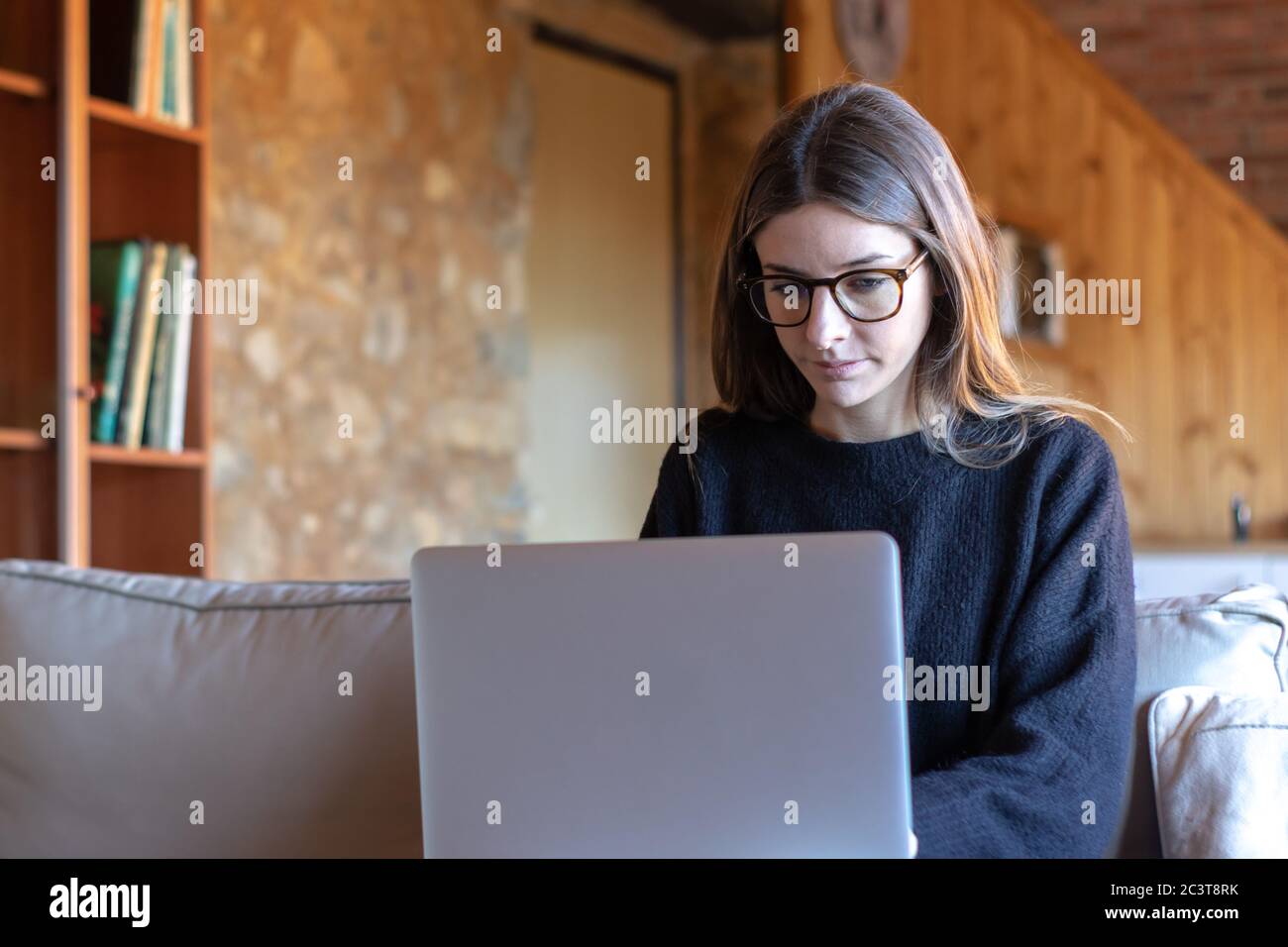 Seriöse junge Frau mit Brille vor dem Laptop schreiben, die zu Hause auf dem Sofa sitzt Stockfoto