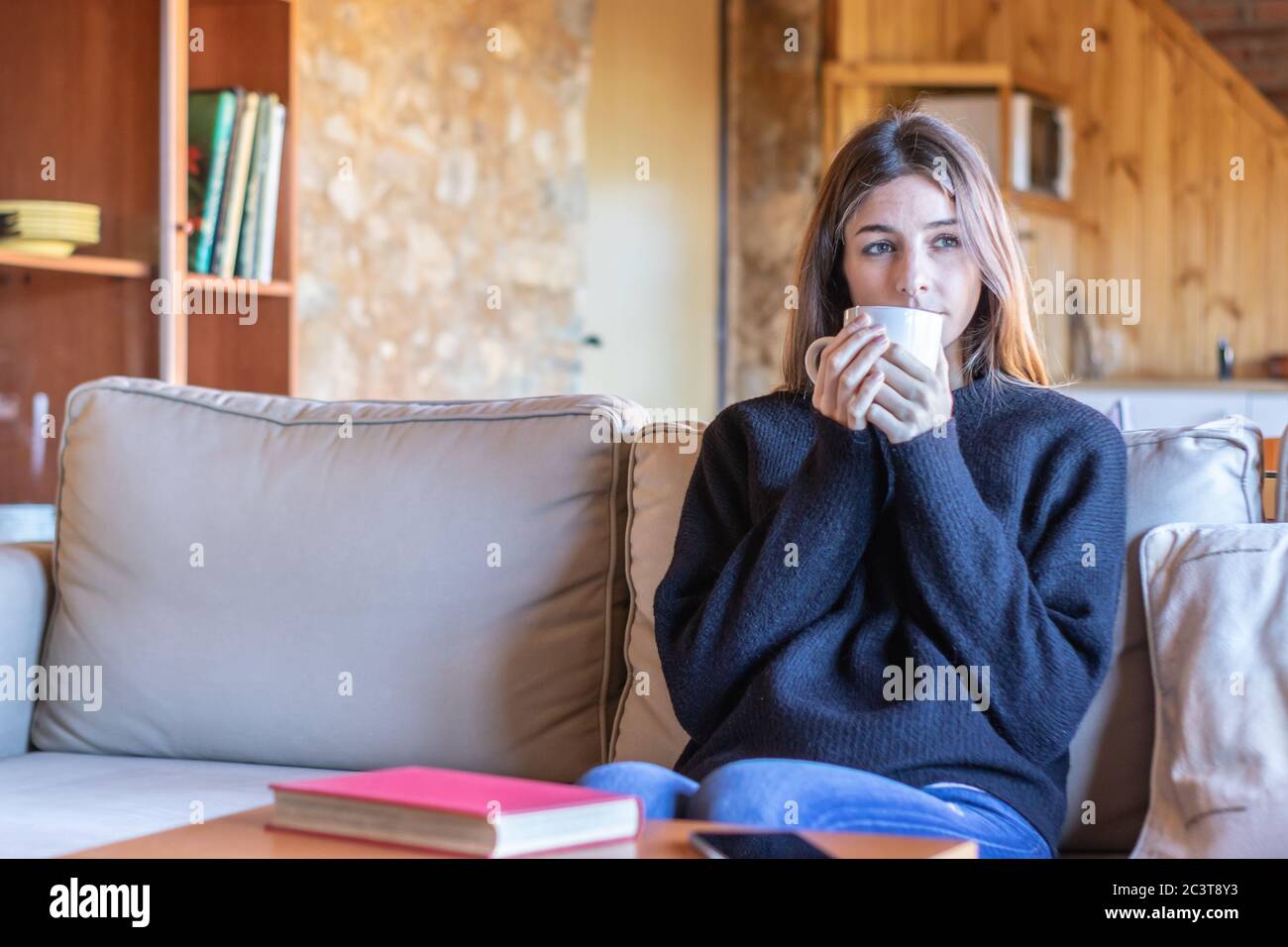 Nostalgische hübsche Mädchen, die Tee trinken sitzen auf dem Sofa zu Hause tragen schwarzen Pullover und blaue Jeans Stockfoto