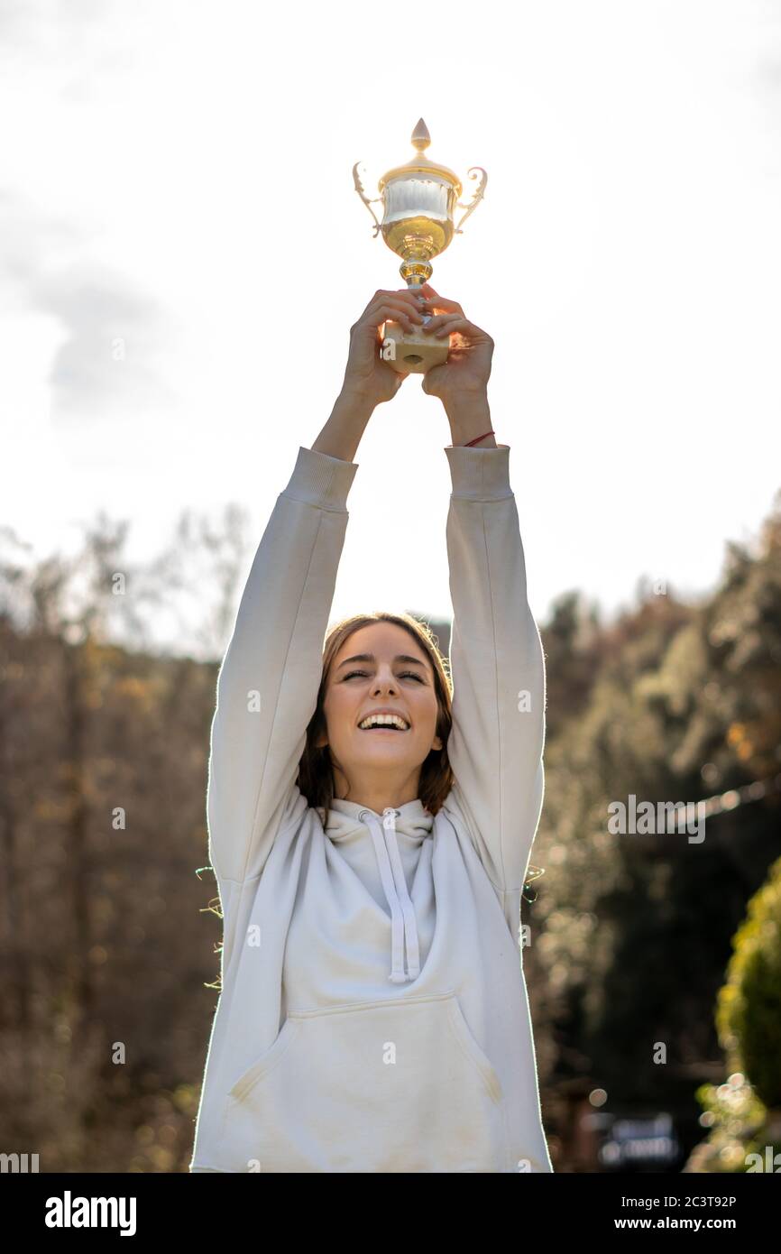 Hübsche lächelnde junge Frau hält eine Trophäe im Freien in einem Park. Sie trägt ein leichtes Sweatshirt und hat lange braune Haare. Stockfoto