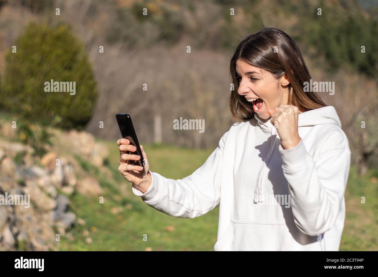 Schöne euphorische Brünette Frau liest Nachrichten auf dem Smartphone in einem Park an einem sonnigen Tag. Sie trägt einen weißen Pullover und hat lange Haare. Stockfoto