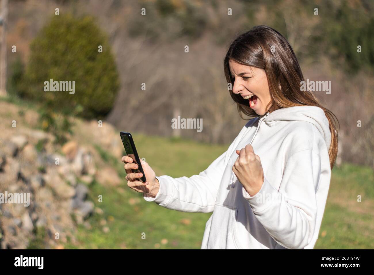 Schöne euphorische Brünette Frau liest Nachrichten auf dem Smartphone in einem Park an einem sonnigen Tag. Sie trägt einen weißen Pullover und hat lange Haare. Stockfoto