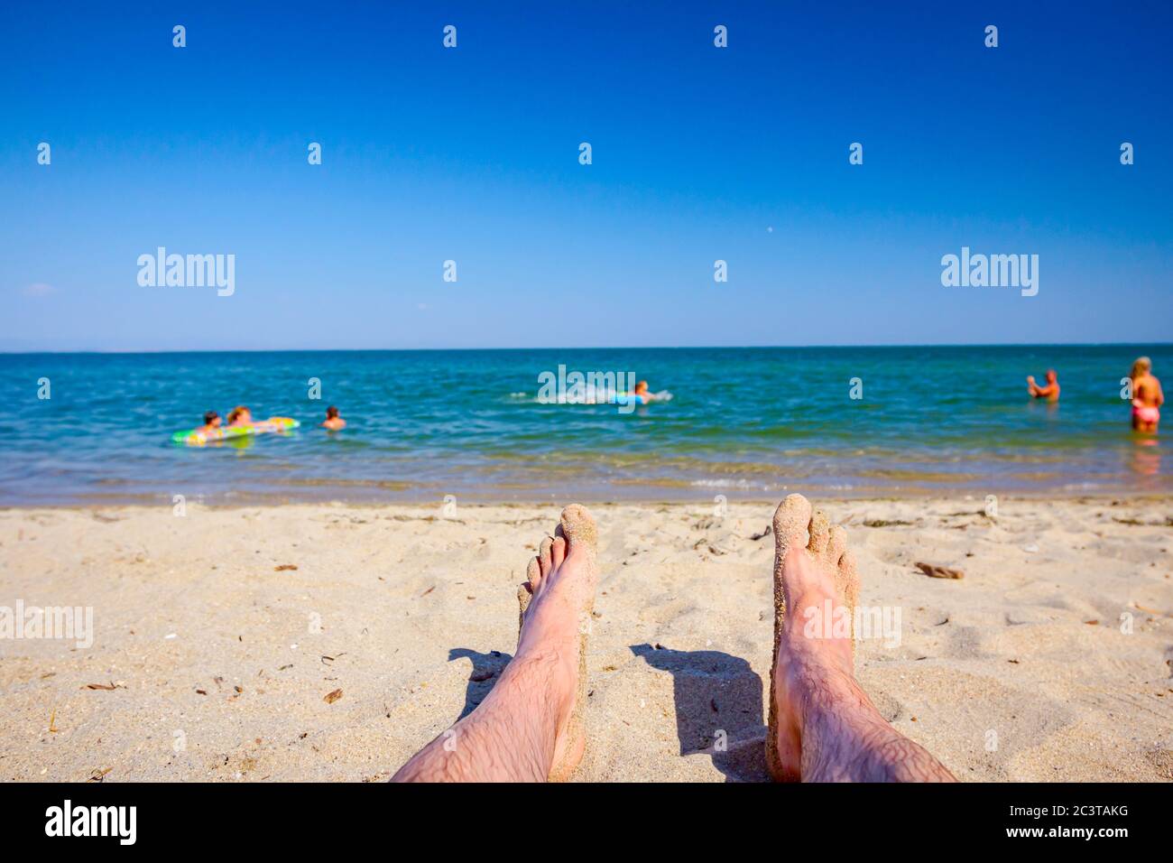 Die Beine des Mannes sonnen sich, indem sie sorglos auf Sand neben der Küste liegen, am öffentlichen Strand. Stockfoto