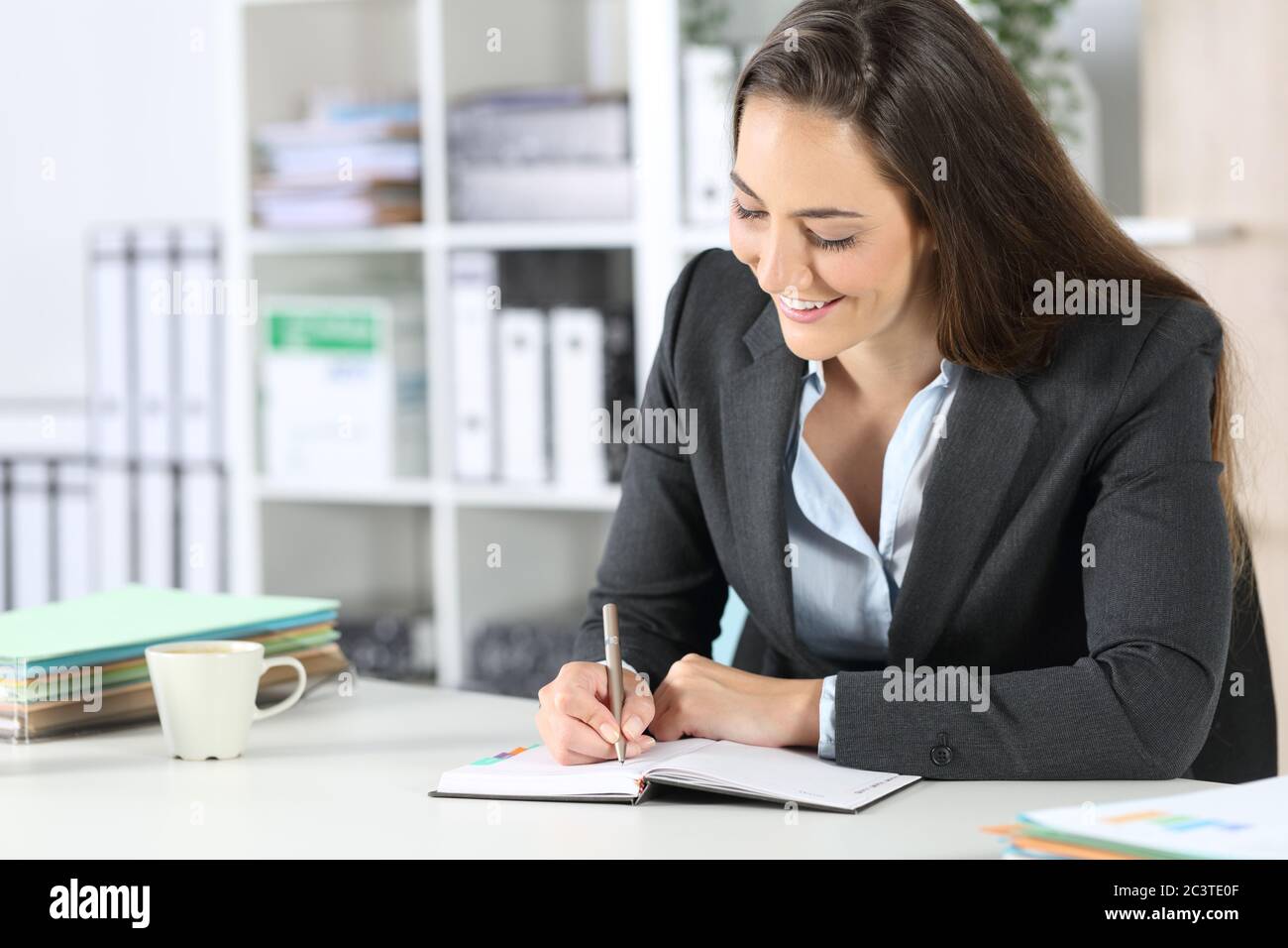 Glückliche Geschäftsführerin, die Notizen auf die Tagesordnung schrieb, die auf einem Schreibtisch im Büro saß Stockfoto