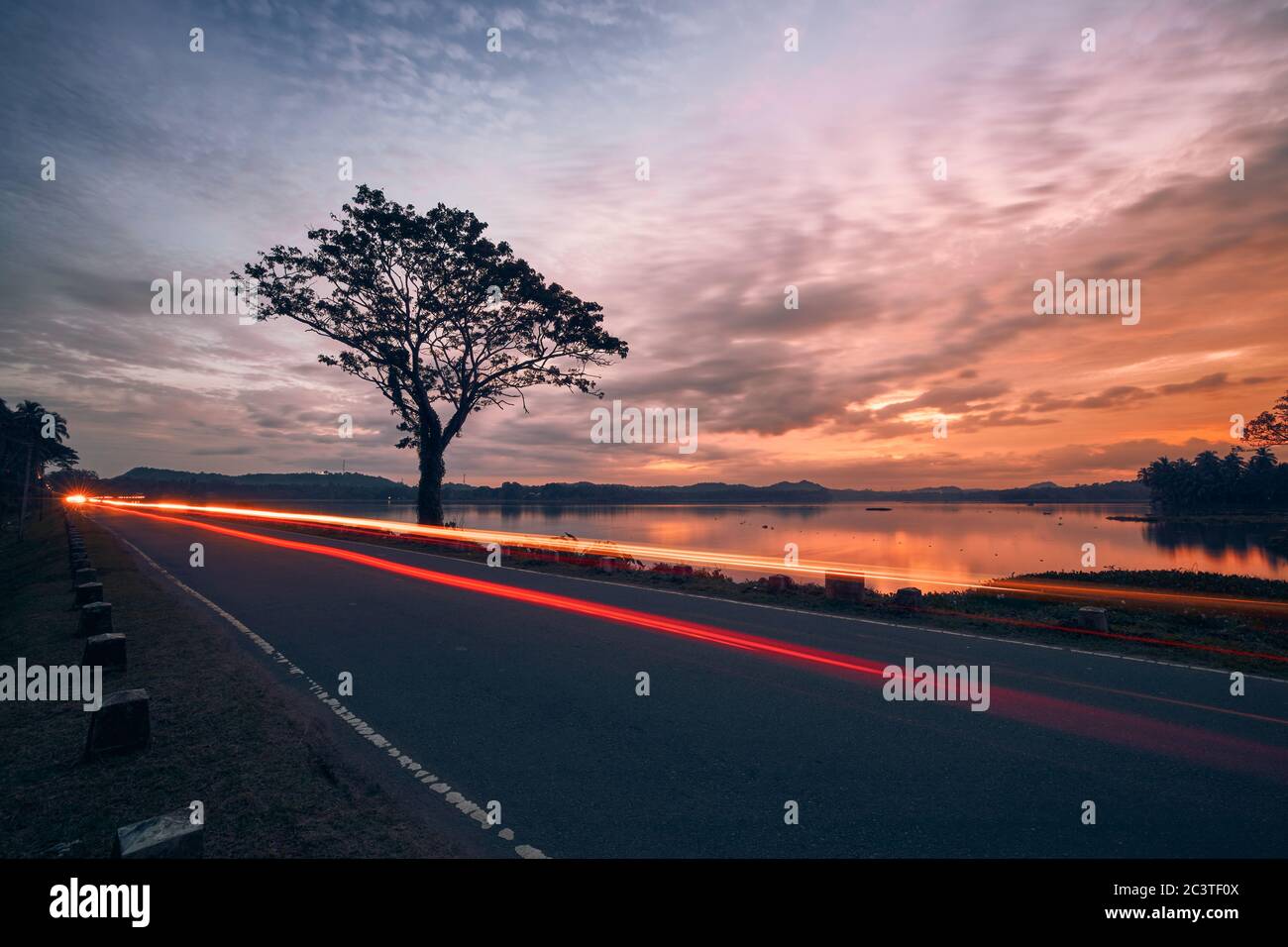 Leichte Autospuren auf der Straße gegen den See bei schönem Sonnenuntergang. Abendverkehr in der Landschaft von Sri Lanka. Stockfoto