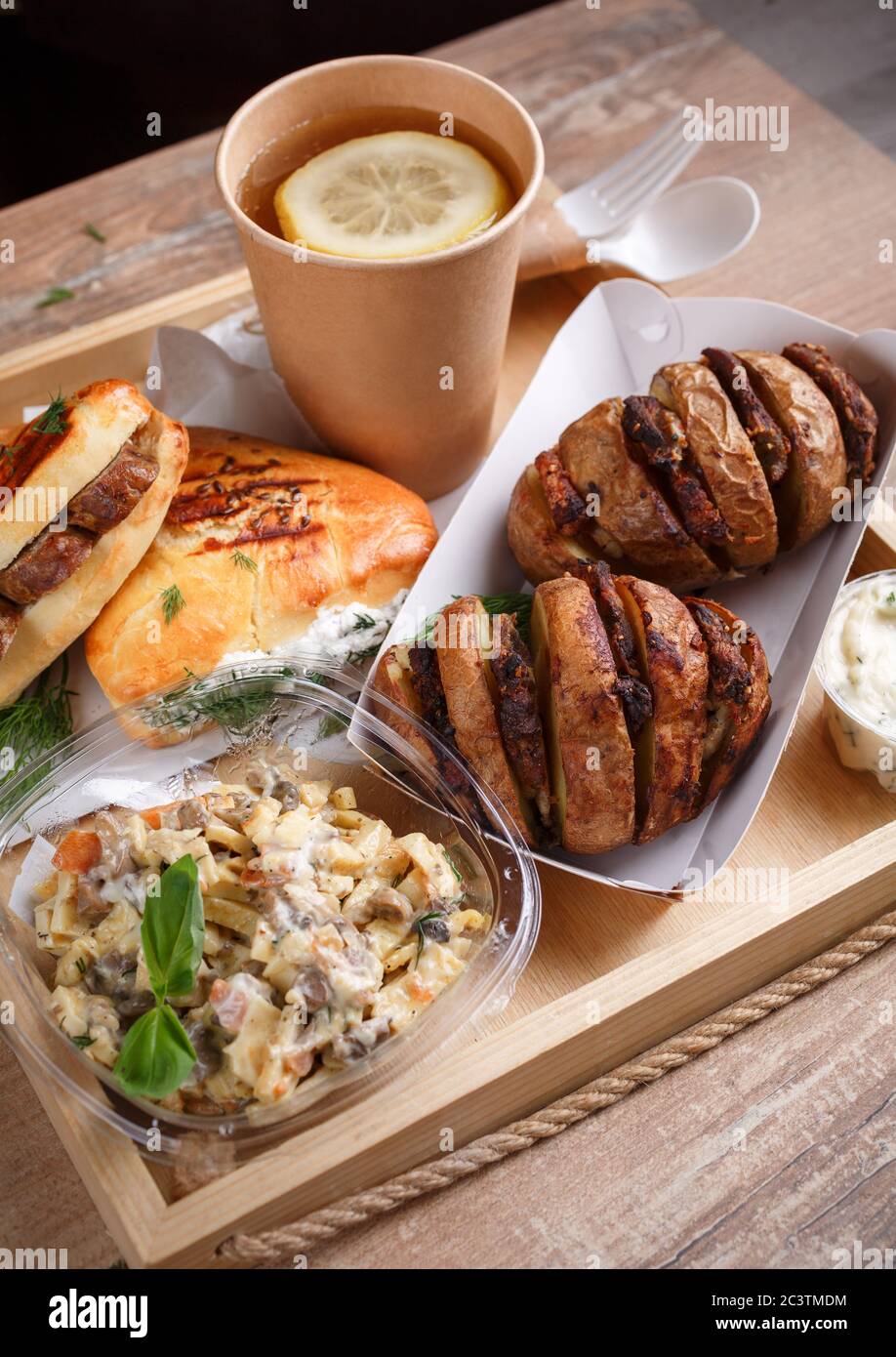 Einweggeschirr, Zitronentee, Schiefer, leckere Wurstbrötchen und Dill und gebackene Kartoffeln mit Fleisch auf einem Holztablett Seitenansicht. Stockfoto