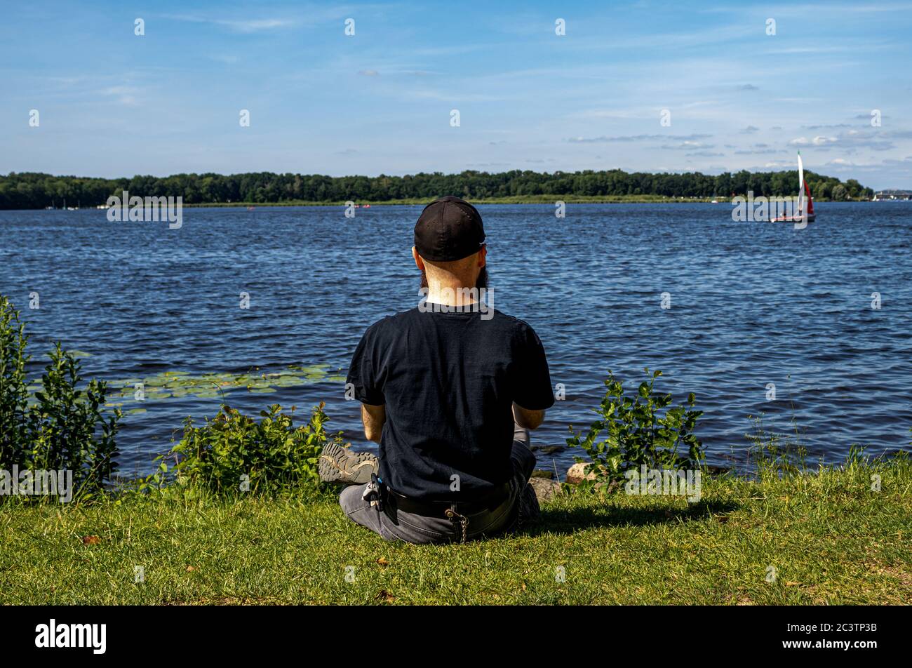 Ein einbärtiger Mann sitzt auf dem grünen Gras auf dem See und schaut auf das Boot. Stockfoto
