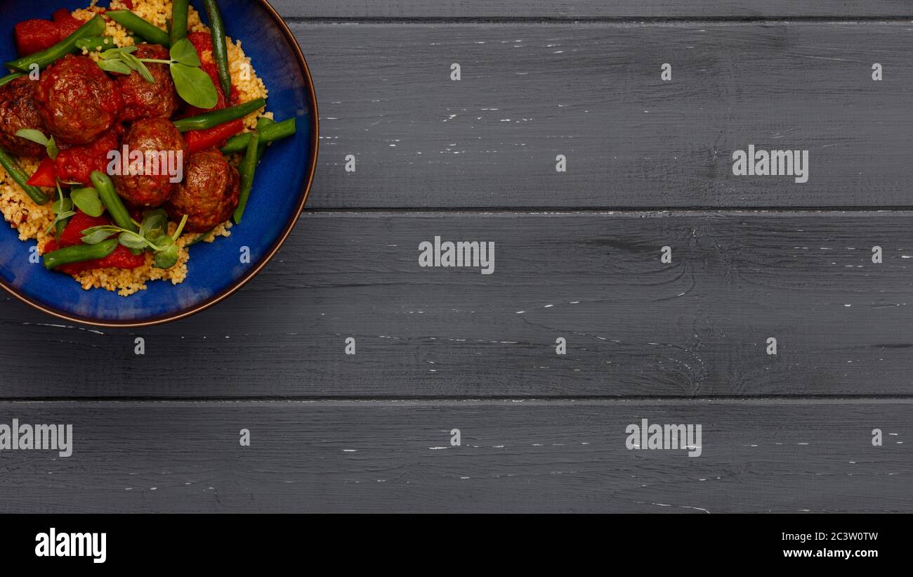 Eine köstliche Schüssel mit Hackfleischbällchen, Couscous und Gemüse, in einer rustikalen blauen Schale auf dunklem Holzhintergrund im Bannerformat Stockfoto
