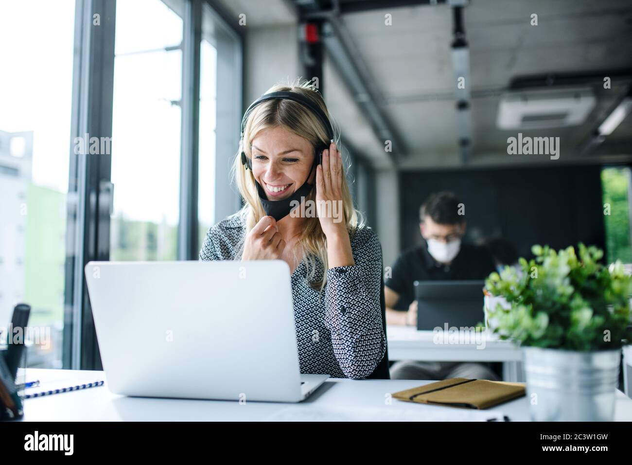 Junge Frau mit Gesichtsmaske und Kopfhörer wieder bei der Arbeit im Büro nach dem Lockdown. Stockfoto