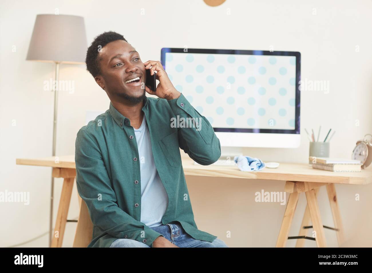 Porträt eines jungen afroamerikanischen Mannes, der per Telefon spricht und lächelt, während er am Schreibtisch in einem modernen Zuhause sitzt, Kopierraum Stockfoto