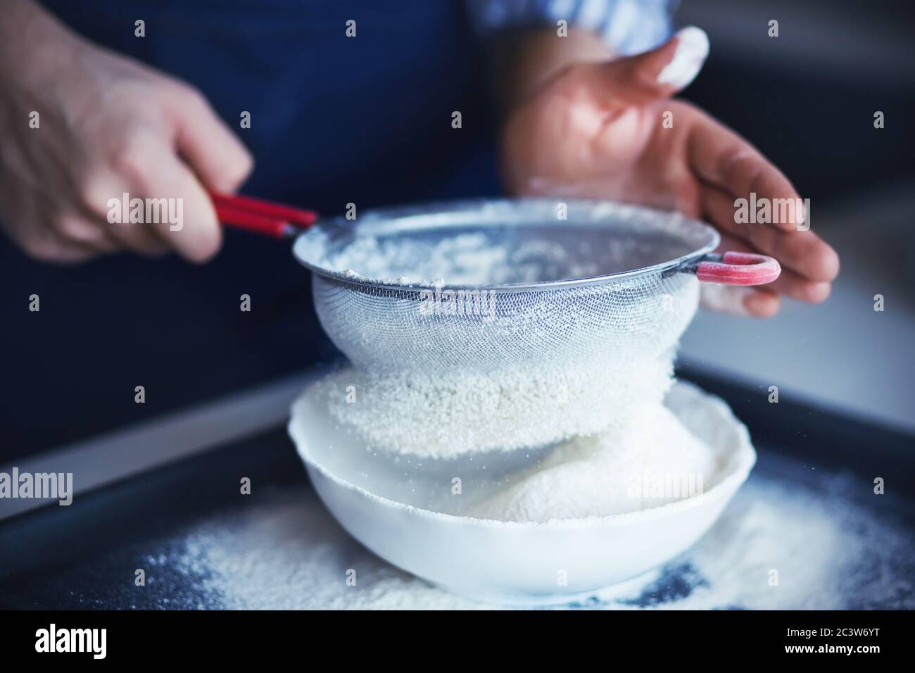 Ein Koch mit seinen Händen, die in Mehl verschmutzt sind, gießt das Mehl durch ein Sieb mit rotem Griff in eine weiße Schüssel. Garvorgang. Stockfoto