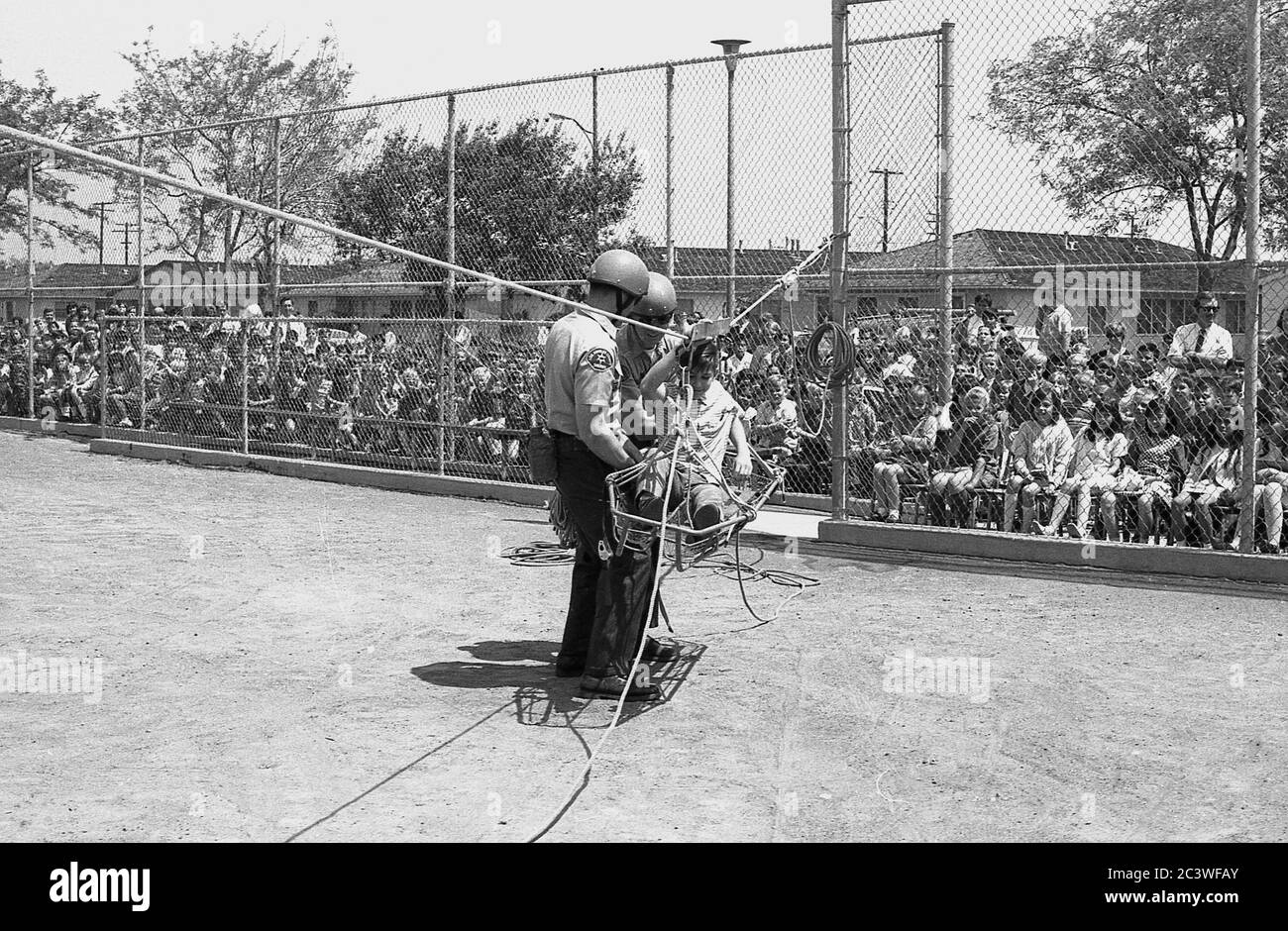 In den 1970er Jahren zeigen zwei Mitglieder der Polizeibehörde VON LA, draußen auf einem Schotterspielplatz, beobachtet von einem Publikum von Schulkindern, wie sie Rettungsaktionen mit Verletzten durchführen, mit Hilfe eines Schuljungen, der in einer Trage und einem an einer Winde befestigten Gurtzeug sitzt, Los Angeles, Kalifornien, USA. Stockfoto