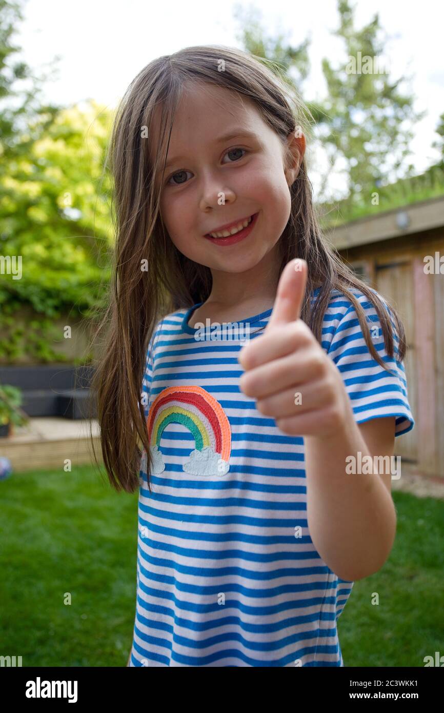 Junge glückliche Mädchen mit Daumen nach oben trägt ein gestreiftes T-Shirt mit Regenbogen Stockfoto