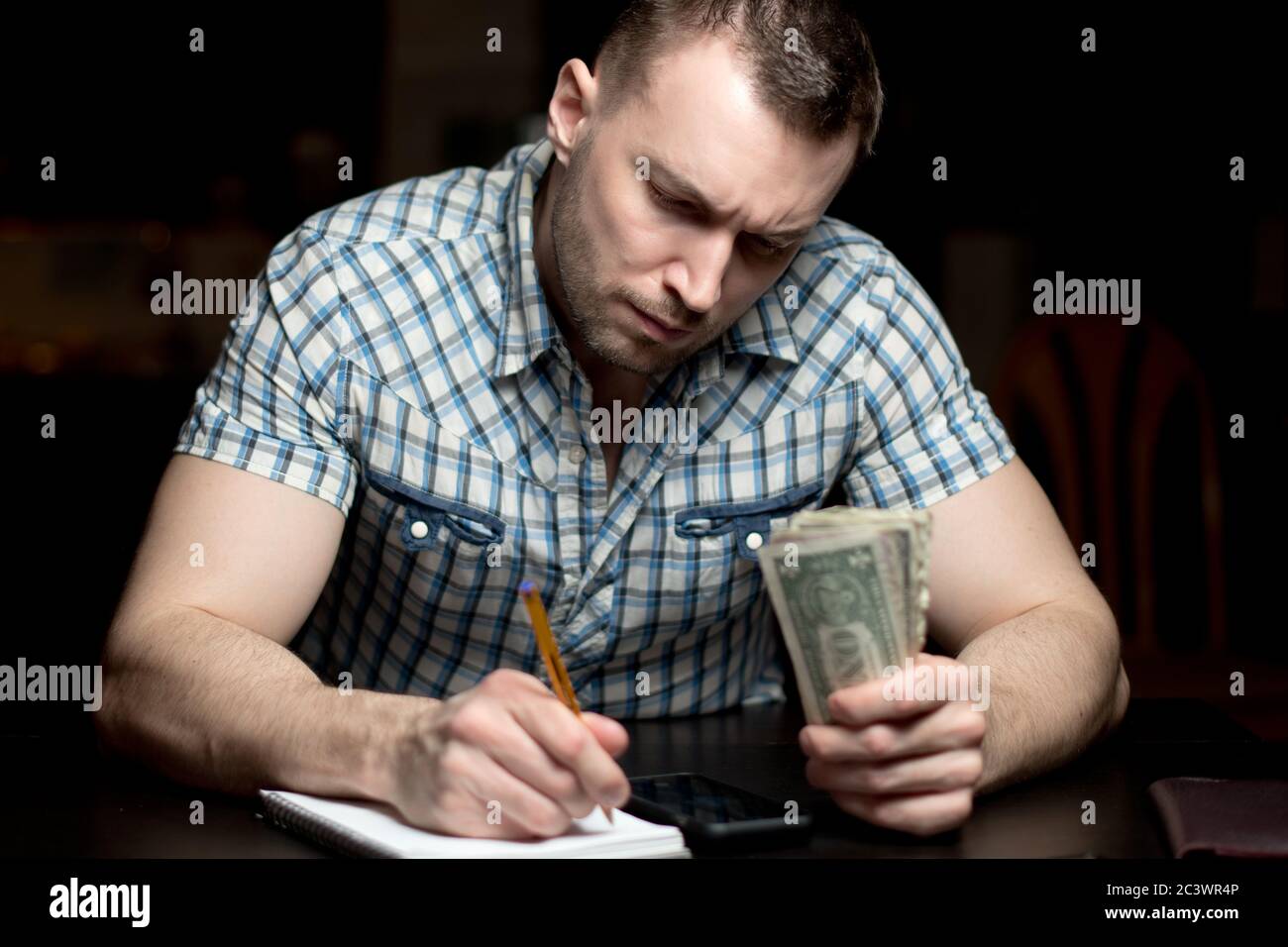Ein Mann mit einem ernsten Gesicht hält Geld und nimmt Notizen in einem Notizbuch Stockfoto