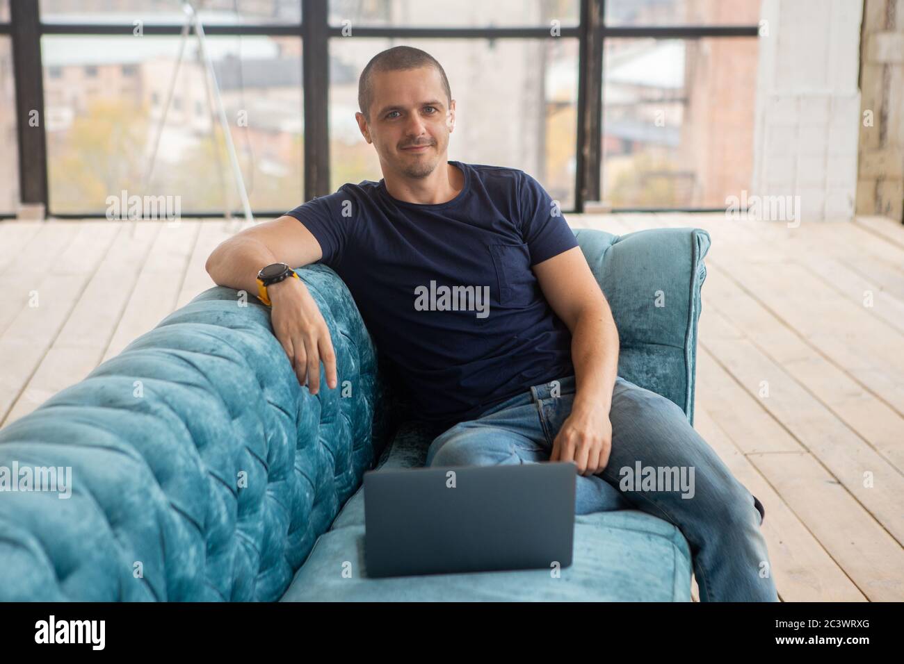 Mann mit Laptop sitzt auf dem Sofa, schaut in die Kamera und lächelt. Stockfoto