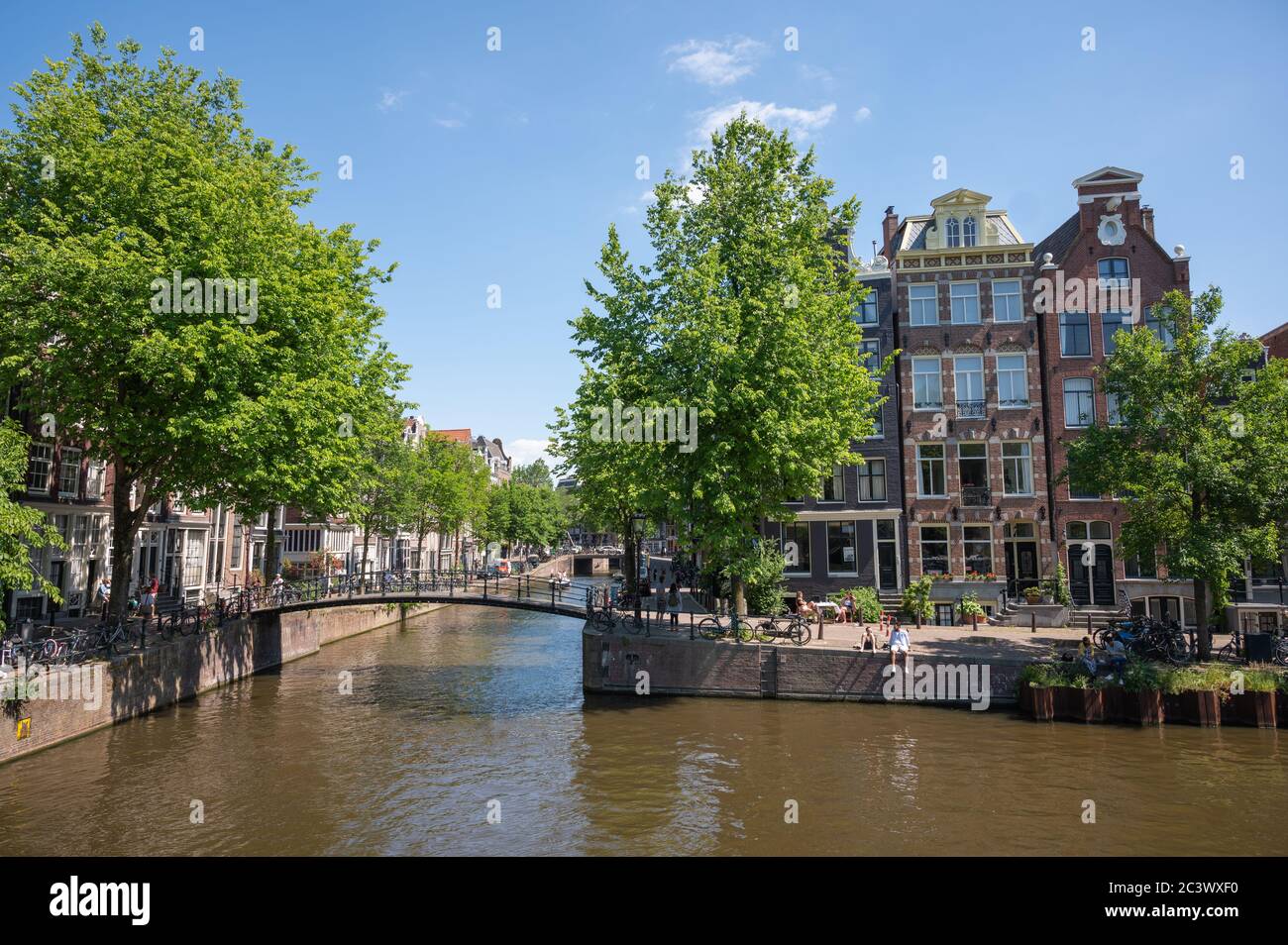 Häuser und Kanal an der Ecke Herengracht und Keizersgracht in Amsterdam, Niederlande. Man sieht Leute sitzen und entspannen auf Kanalseite Stockfoto