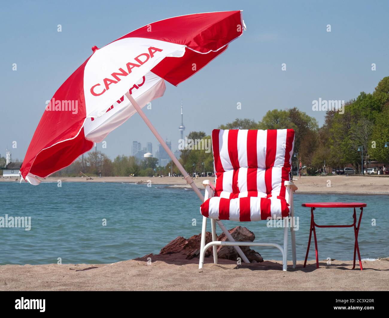 Ein Sonnenschirm mit kanadischer Flagge und ein rot gestreifter Strandstuhl mit der Skyline von Toronto in der Ferne, um den Canada Day am Strand zu feiern Stockfoto