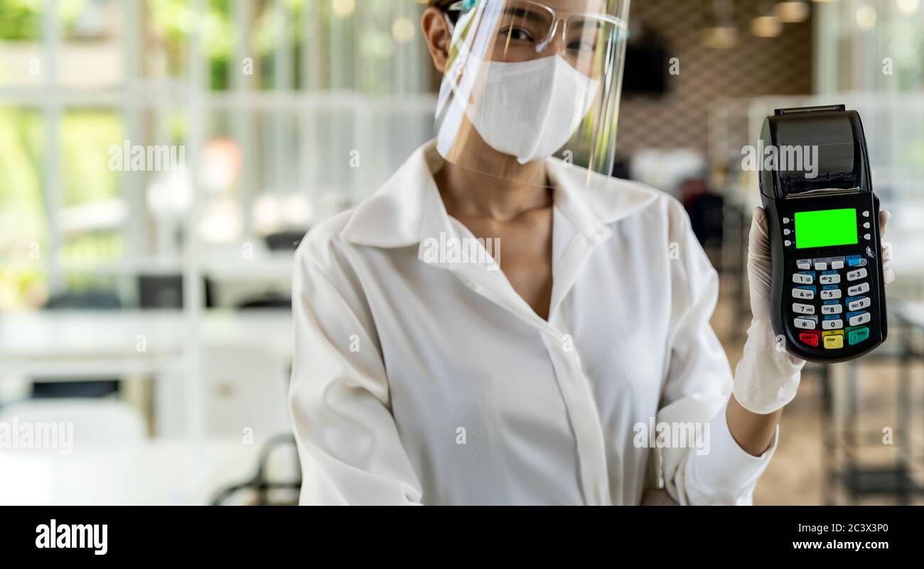 Portrait Attraktive asiatische Kellnerin tragen Gesichtsmaske und Gesichtsschutz halten Kreditkartenleser für kontaktlose Zahlung mit Restaurant innen Hintergrund. N Stockfoto