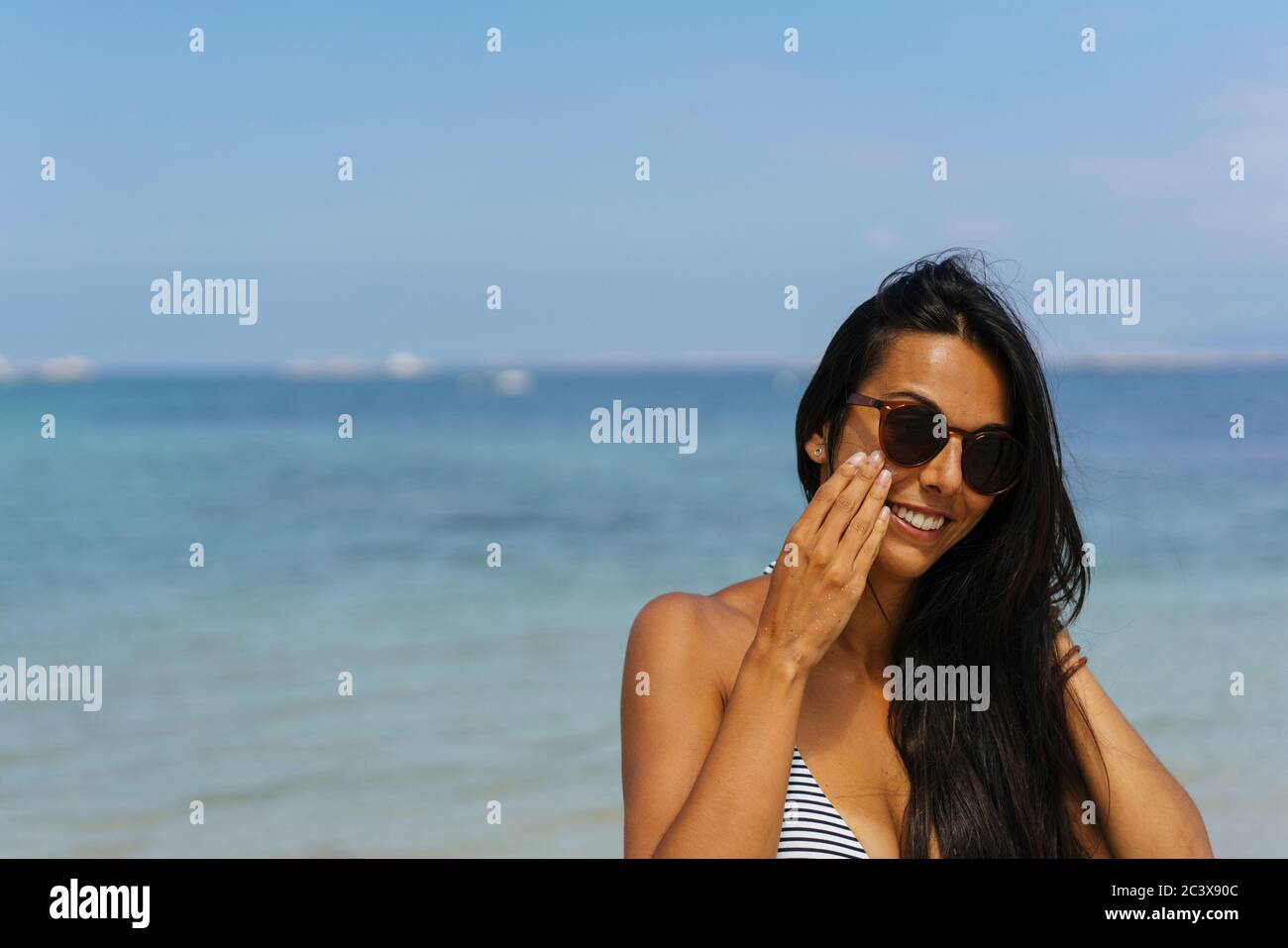 Lächelnde Frau, die Sonnenschutzcreme auf ihr Gesicht am Strand aufgibt Stockfoto