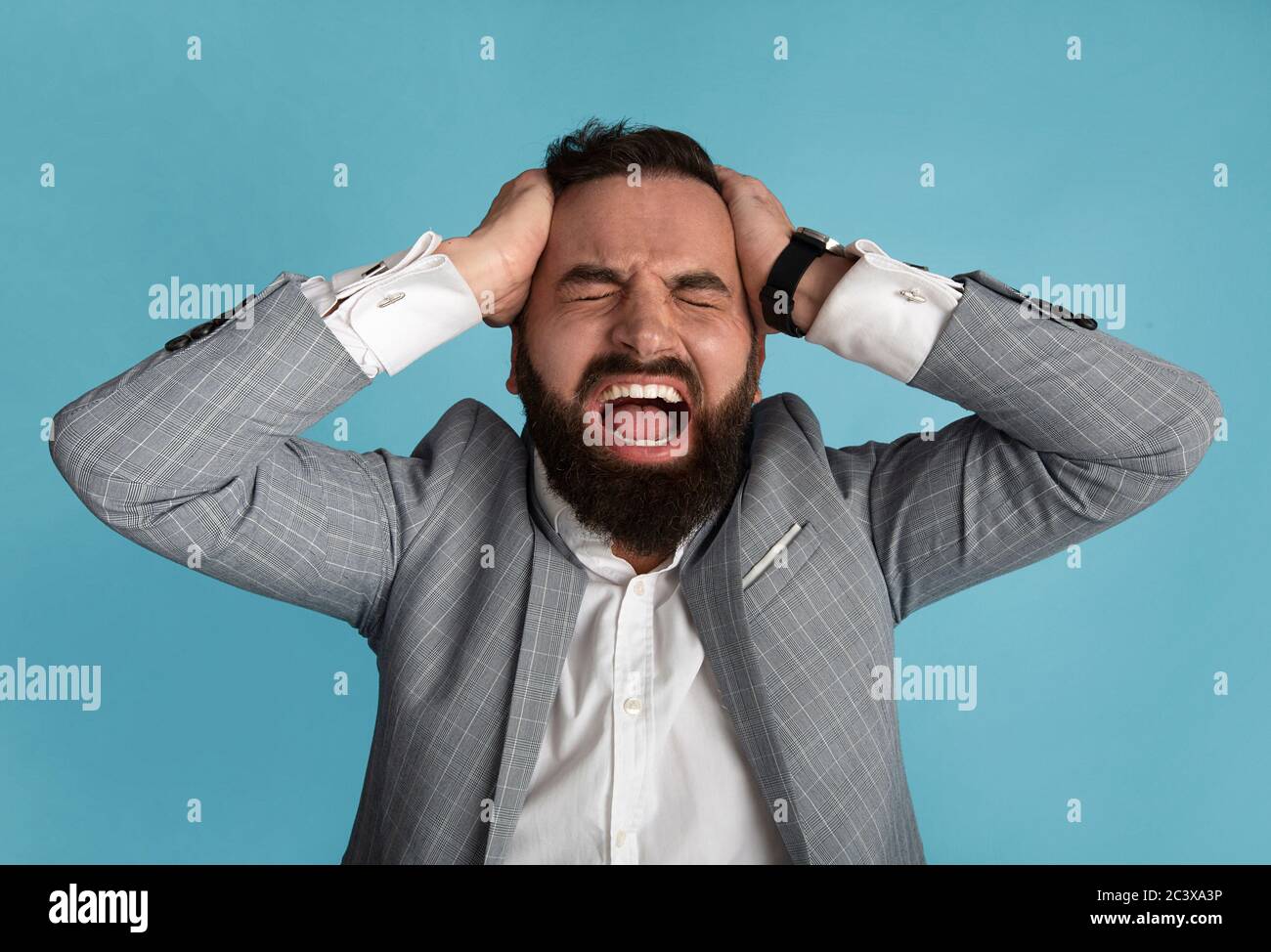 Millennial Geschäftsmann hält seinen Kopf und schreit vor Wut auf blauem Hintergrund Stockfoto