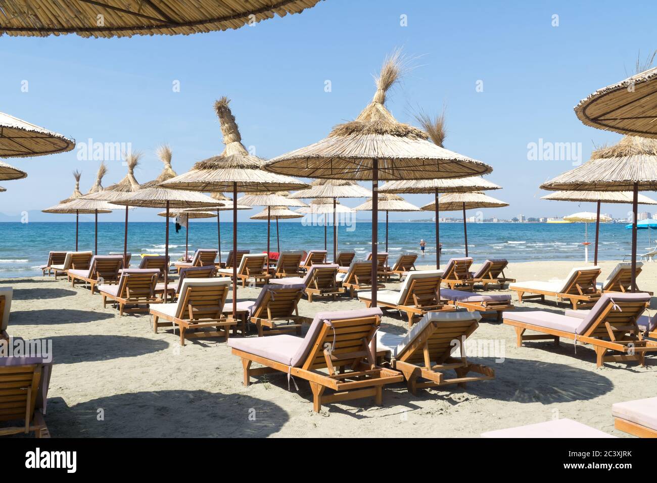 Die Reihen der leeren Liegen mit Strohschirmen am Strand. Blauer Himmel und türkisfarbenes Meer. Sonniger Sommertag. Vlora / Vlore, Albanien, Europa. Stockfoto