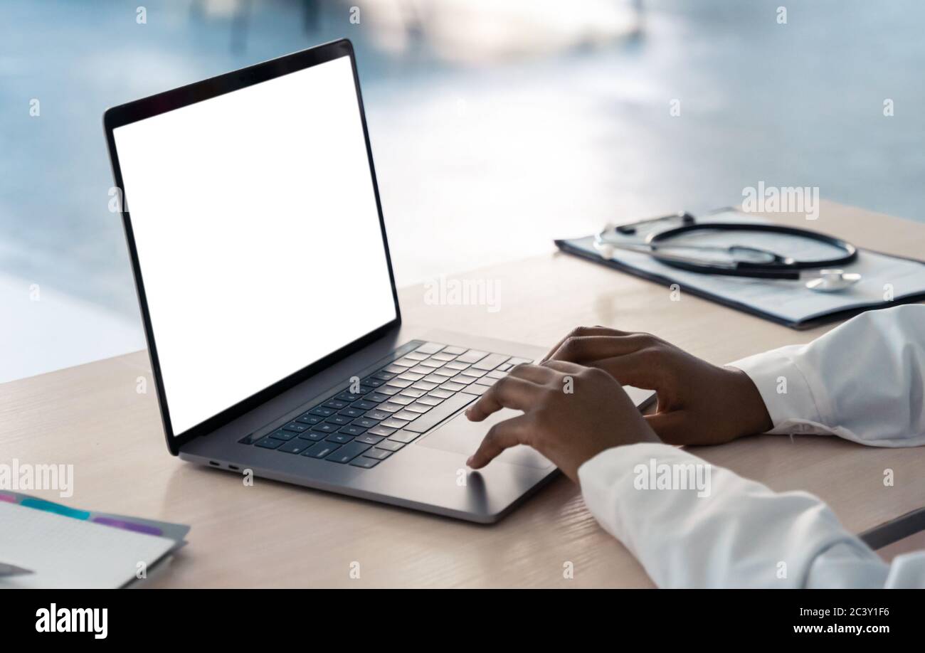 Afrikanischer Arzt mit Laptop tippen Modell weißen Bildschirm am Schreibtisch, Nahaufnahme. Stockfoto