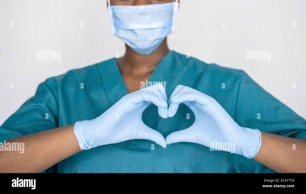 Afrikanische Krankenschwester tragen Gesichtsmaske, Handschuhe, blaue Uniform zeigt Herz Hände Form. Stockfoto