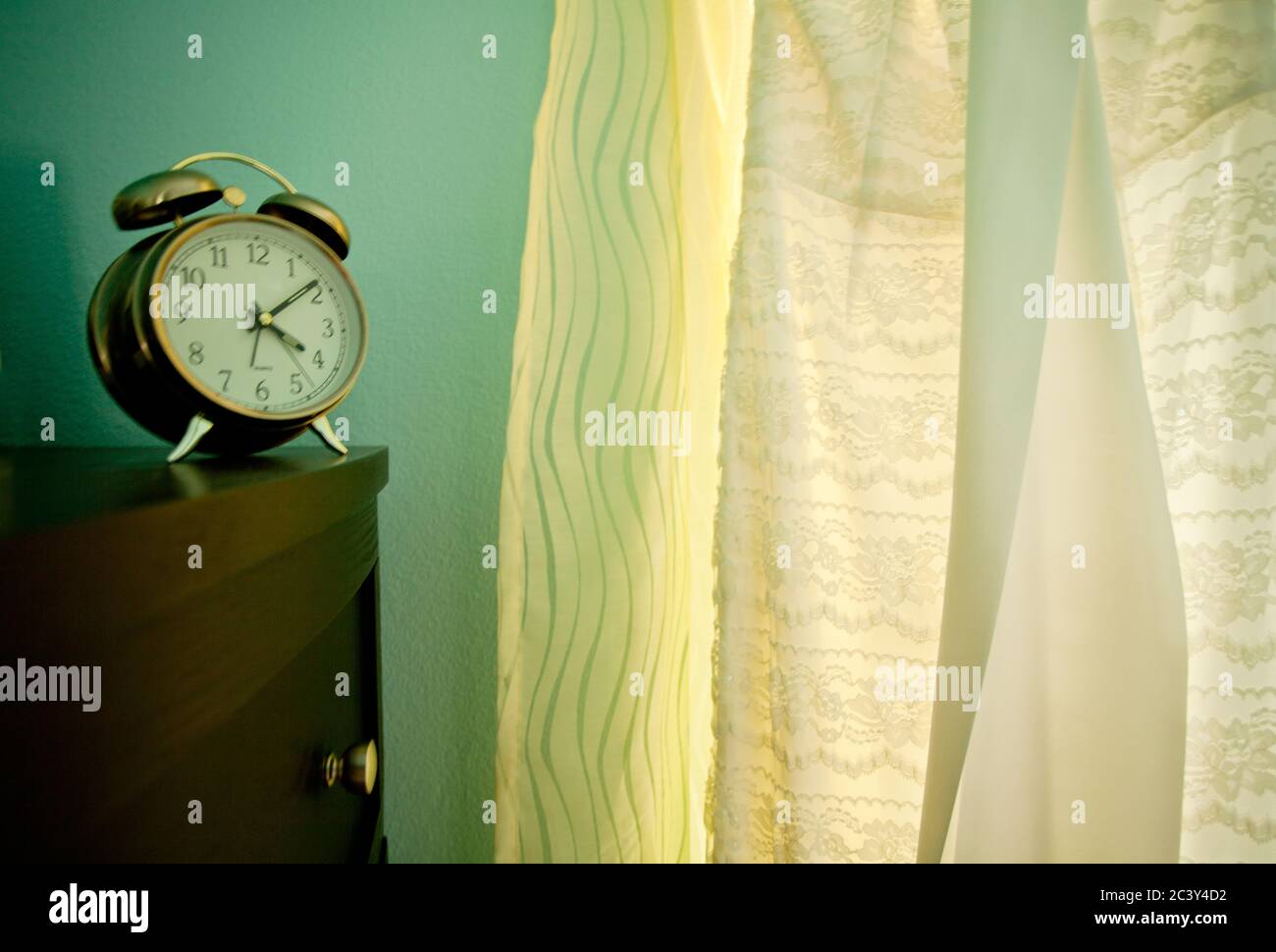 Ein traditioneller Wecker auf einer Kommode neben einem weißen Hochzeitskleid vor einem Fenster. Stockfoto