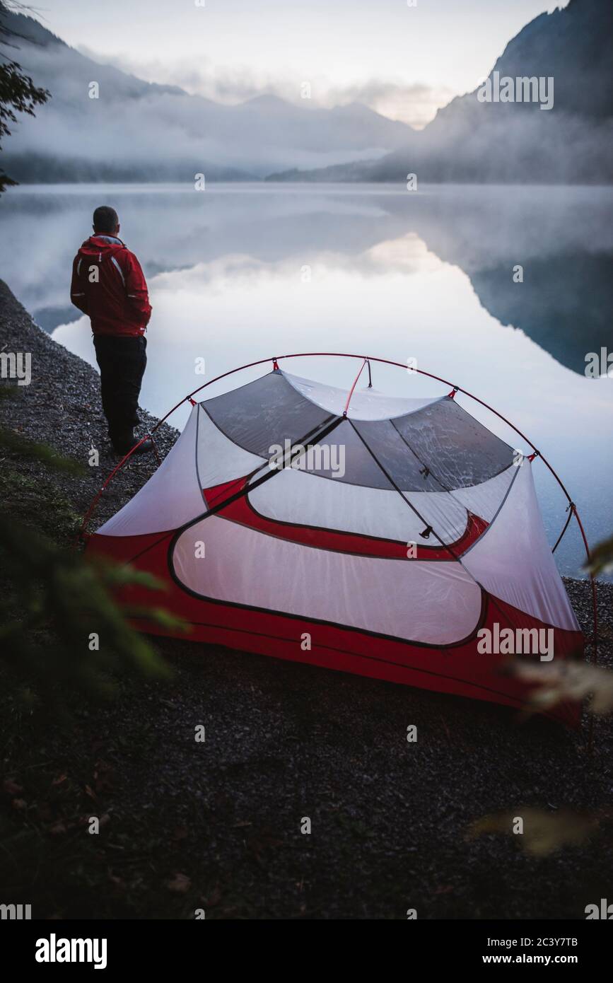 Österreich, Plansee, junger Mann, der in der Nähe des Zeltes steht und den Plansee bei Sonnenaufgang betrachtet Stockfoto