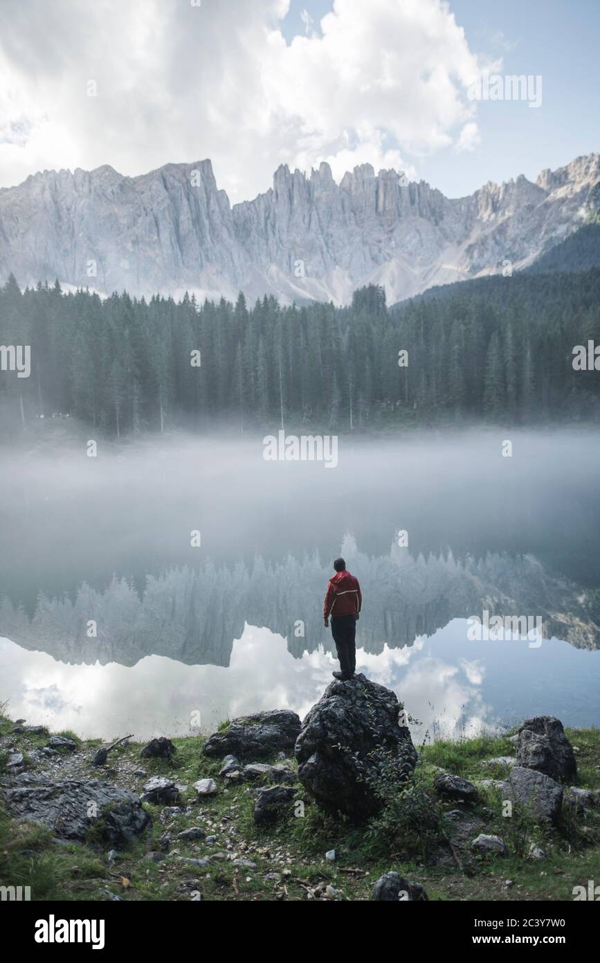 Italien, Karersee, junger Mann steht am Karersee in den Dolomiten bei Sonnenaufgang auf dem Felsen Stockfoto